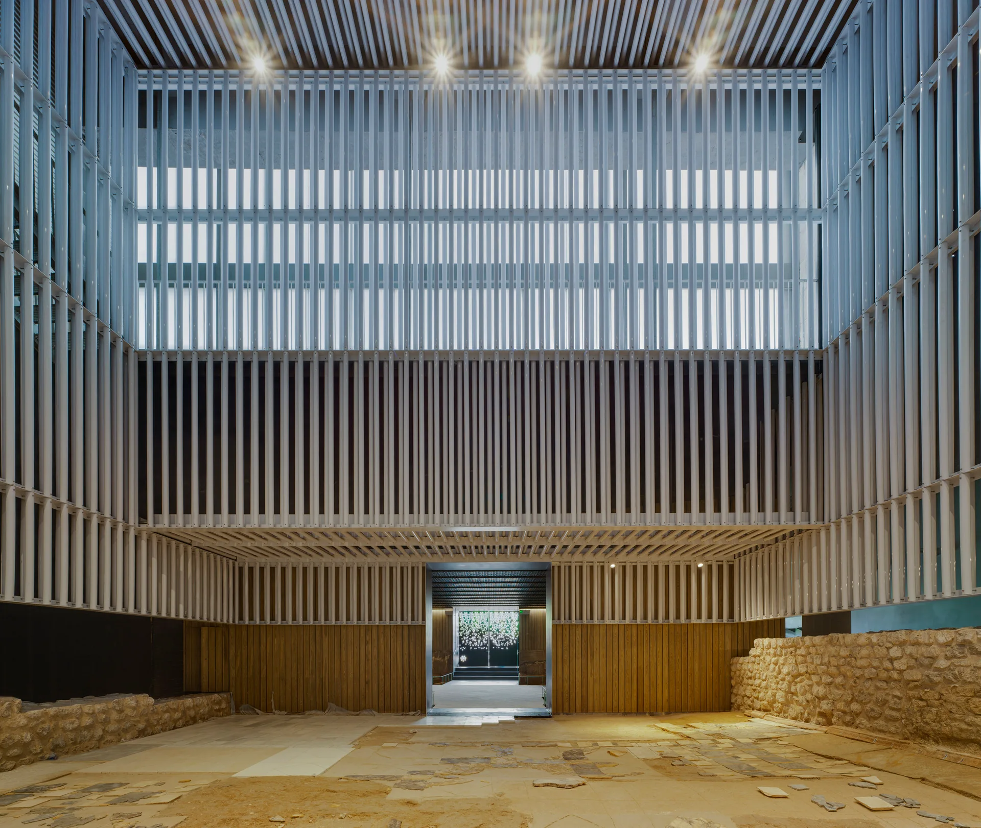 A spacious atrium features soaring white vertical slat walls over warm wood paneling, historic-style rough stone base walls, and a stone slab floor.