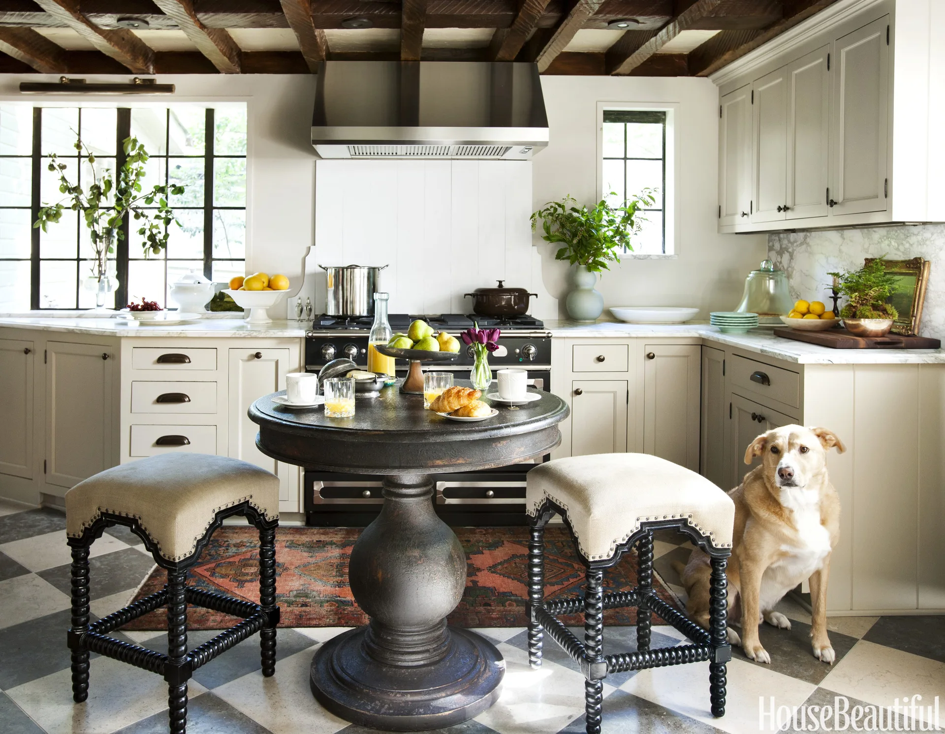 A kitchen features a round dark wood table with upholstered stools, checkered stone tile flooring, painted cabinets, and dark exposed ceiling beams.
