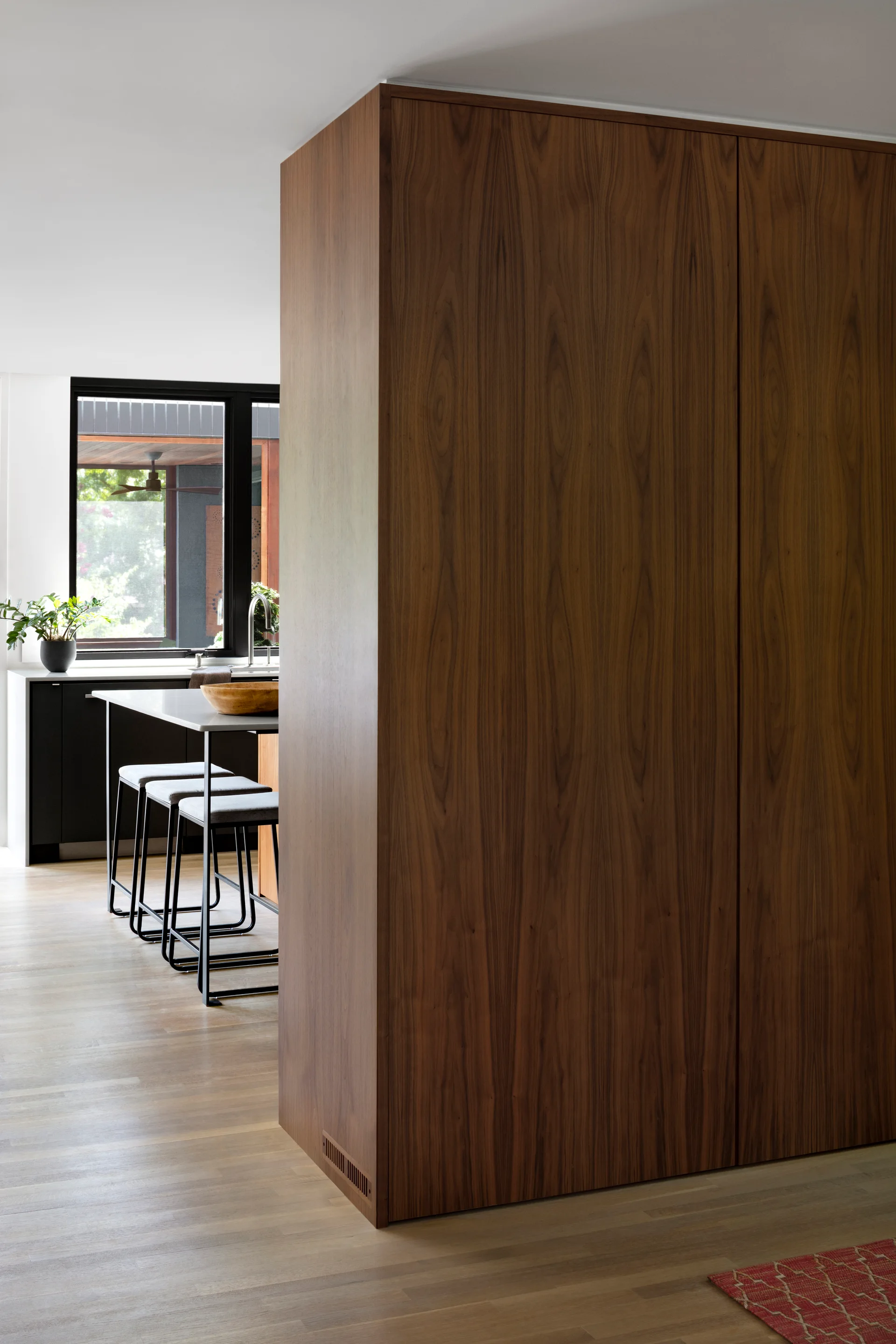 A minimalist kitchen features a large wood grain cabinet unit next to an island with gray bar stools and a black-framed window.