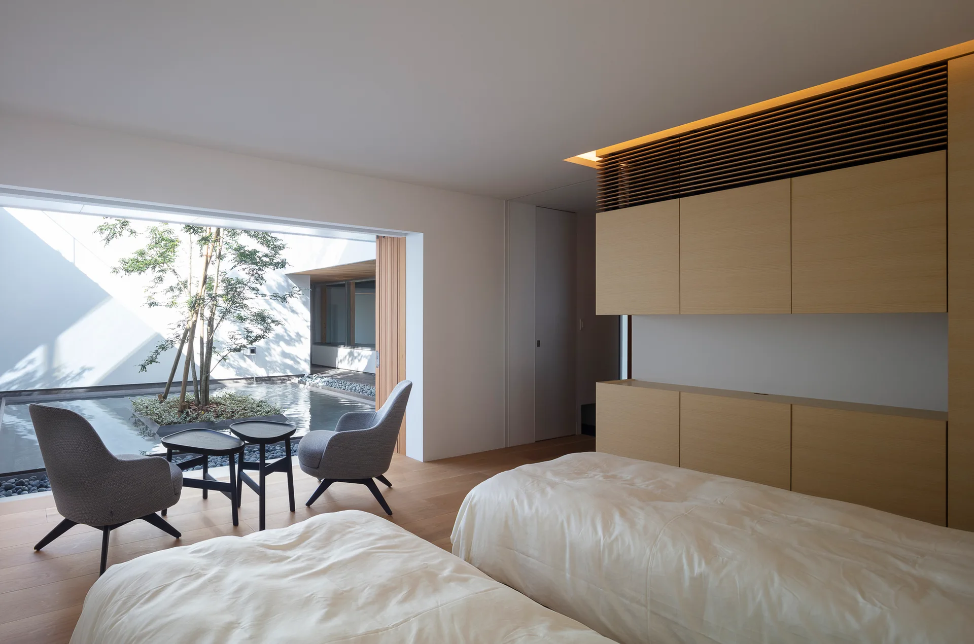 A minimalist bedroom with two beds and gray armchairs faces a sliding glass door opening to a private courtyard pool.
