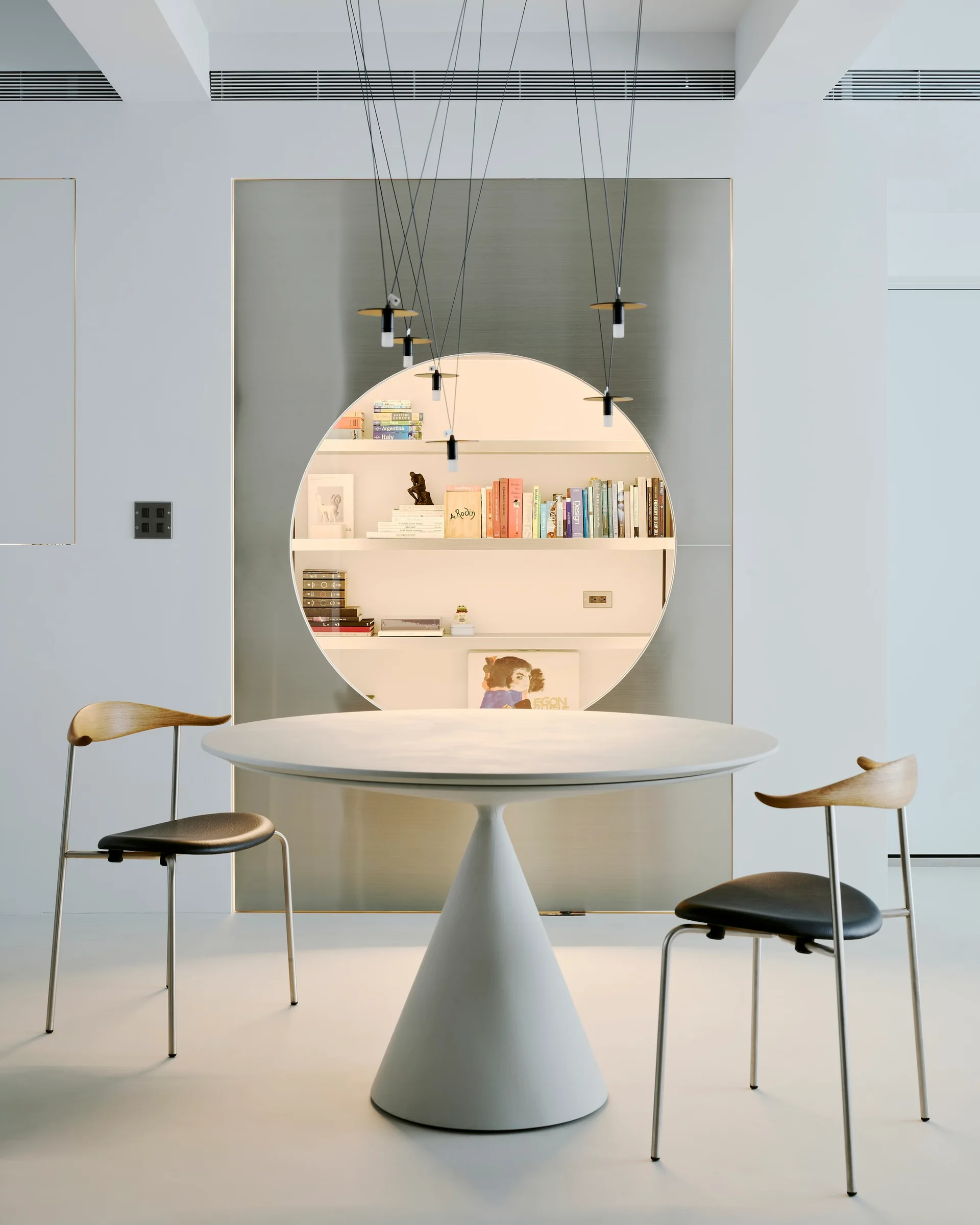 A modern white pedestal dining table sits in front of a brushed metal wall panel with a large, illuminated circular recessed bookshelf.