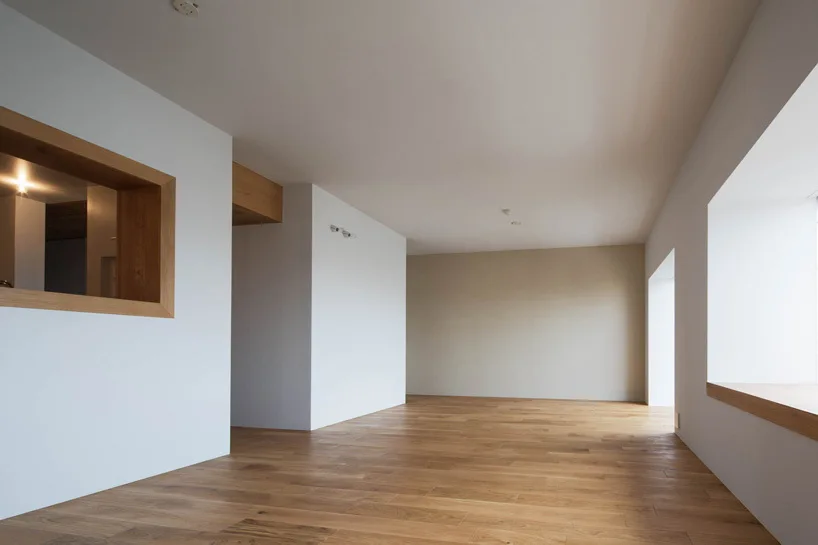 An empty minimalist room features light oak hardwood flooring, plain white walls, a recessed window sill, and a wood-trimmed interior cutout.
