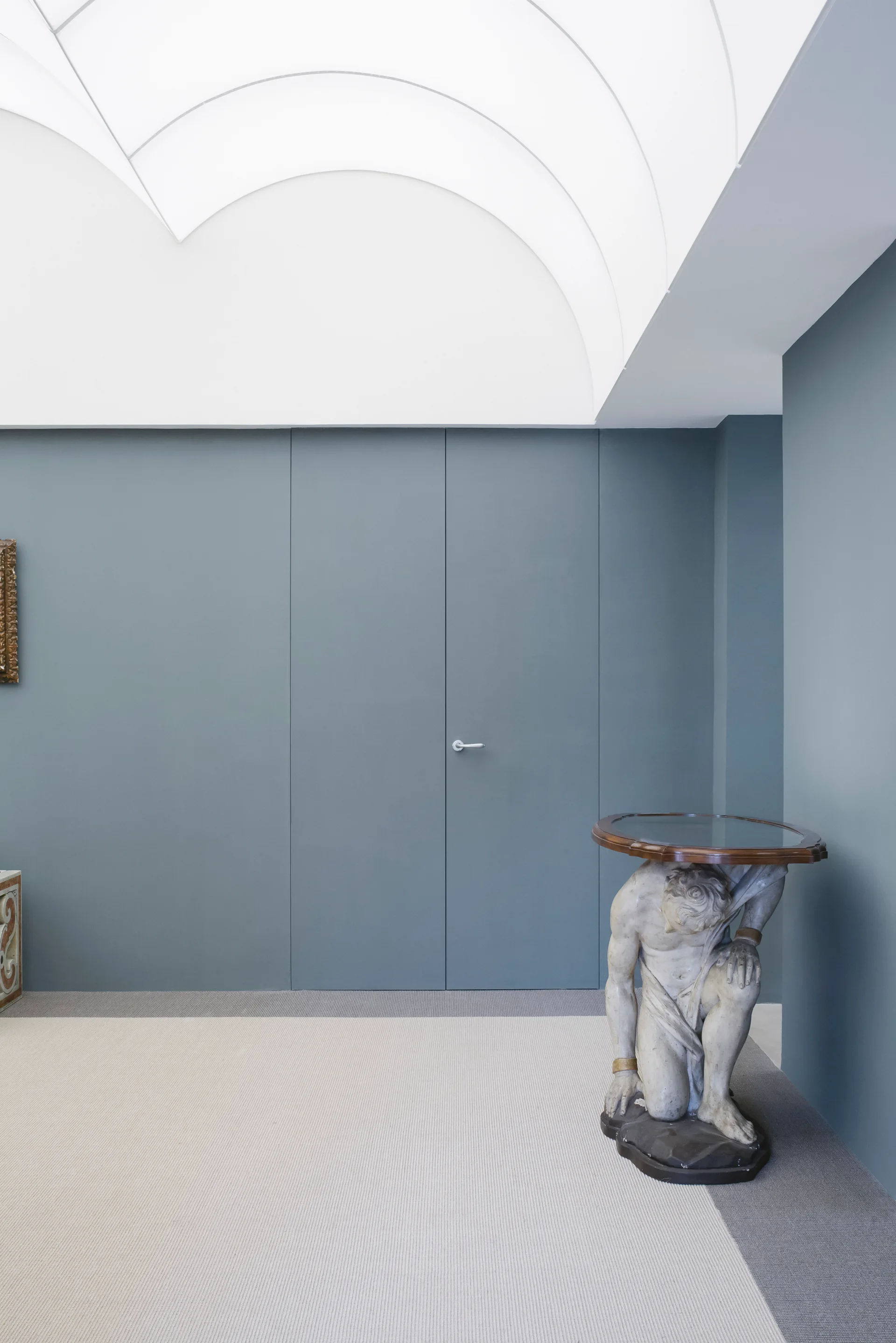 A minimalist room features a slate blue-grey wall with a flush-mounted door, a curved white coved ceiling, and a classical stone-like sculptural side table.