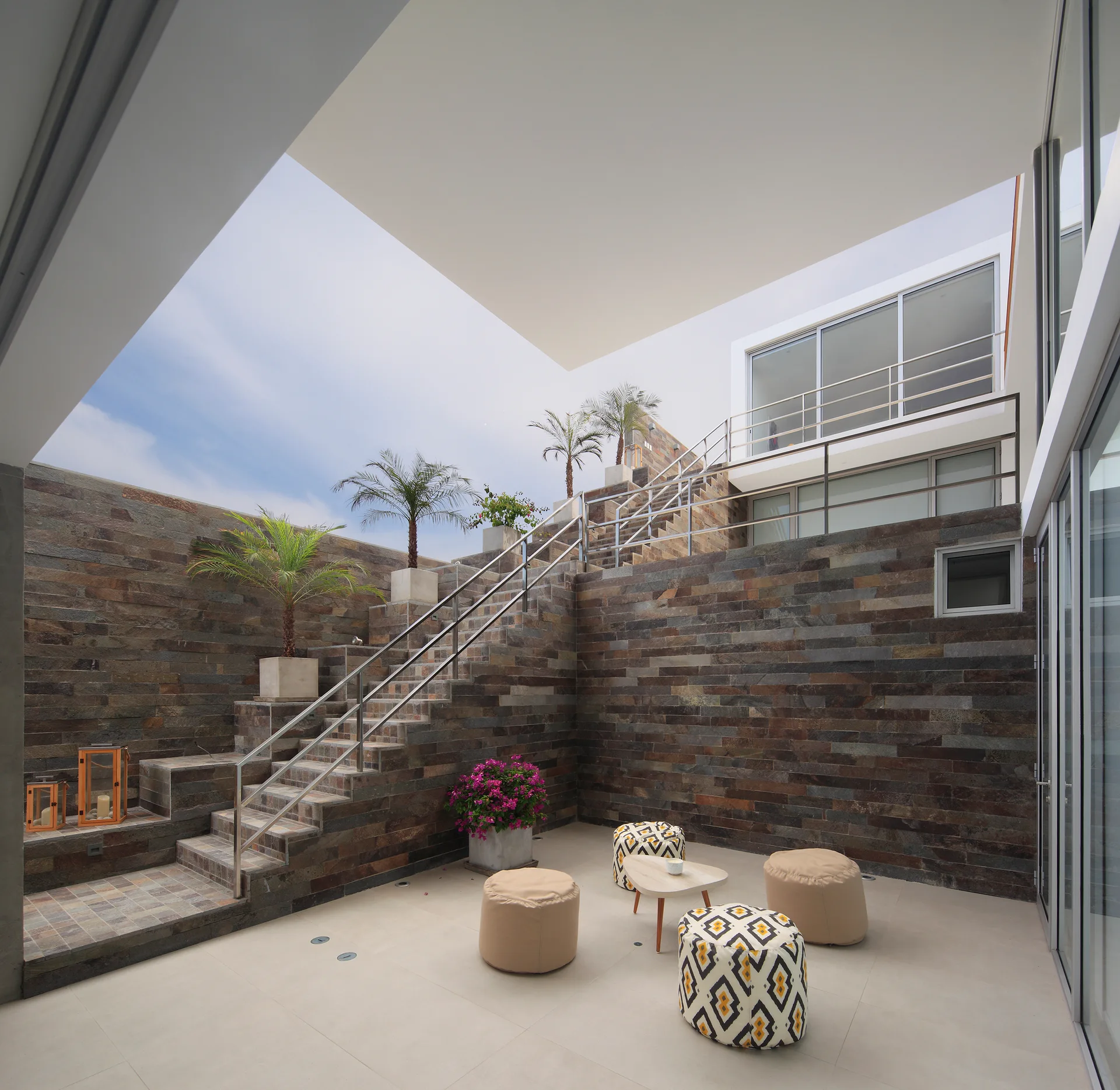 A modern sunken courtyard features high stacked stone walls, a stone staircase with metal handrails, and a seating area with low poufs.