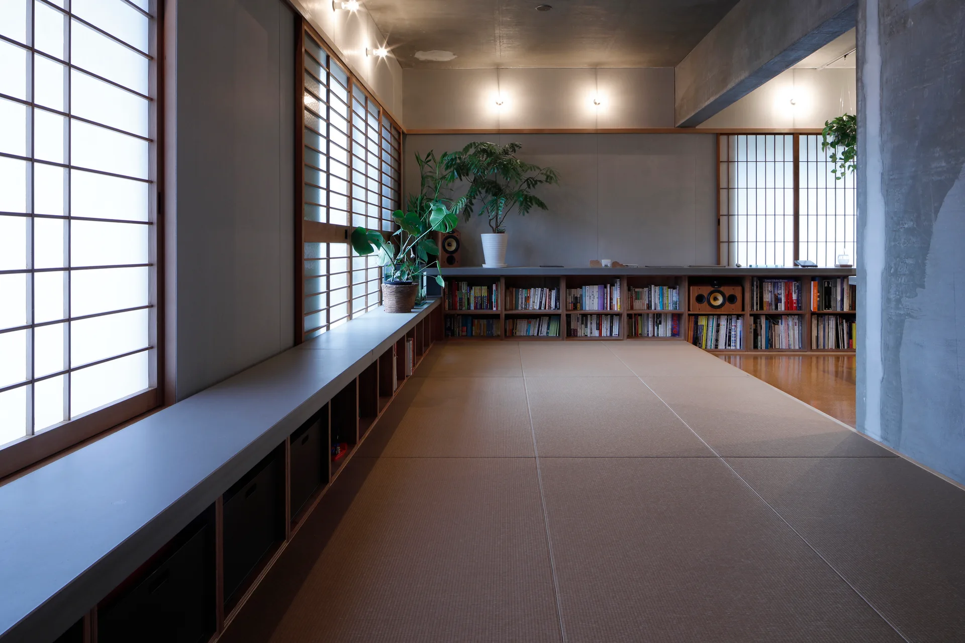 A minimalist Japanese-style room features tan tatami floor mats, raw concrete walls, low built-in wooden bookshelves, and sliding shoji screens over windows.