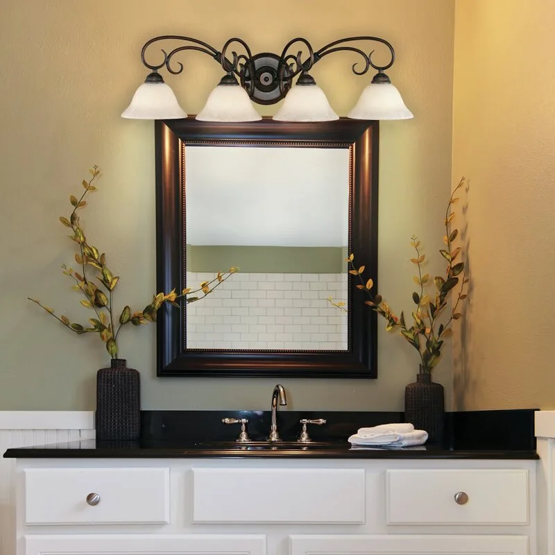 A bathroom vanity features a four-light bronze fixture above a dark-framed mirror, flanked by two dark vases with branches against sage green walls.