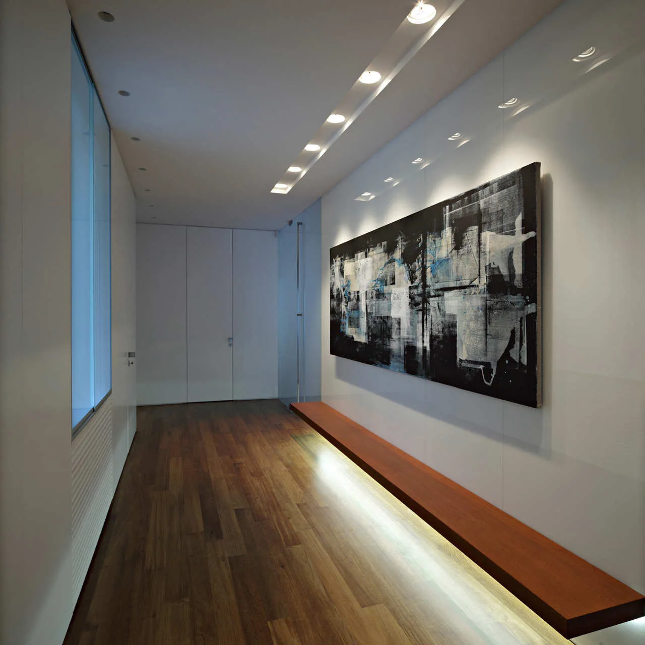 A minimalist hallway features dark wood floors, a long floating bench with warm under-lighting, and a large abstract painting on the wall.