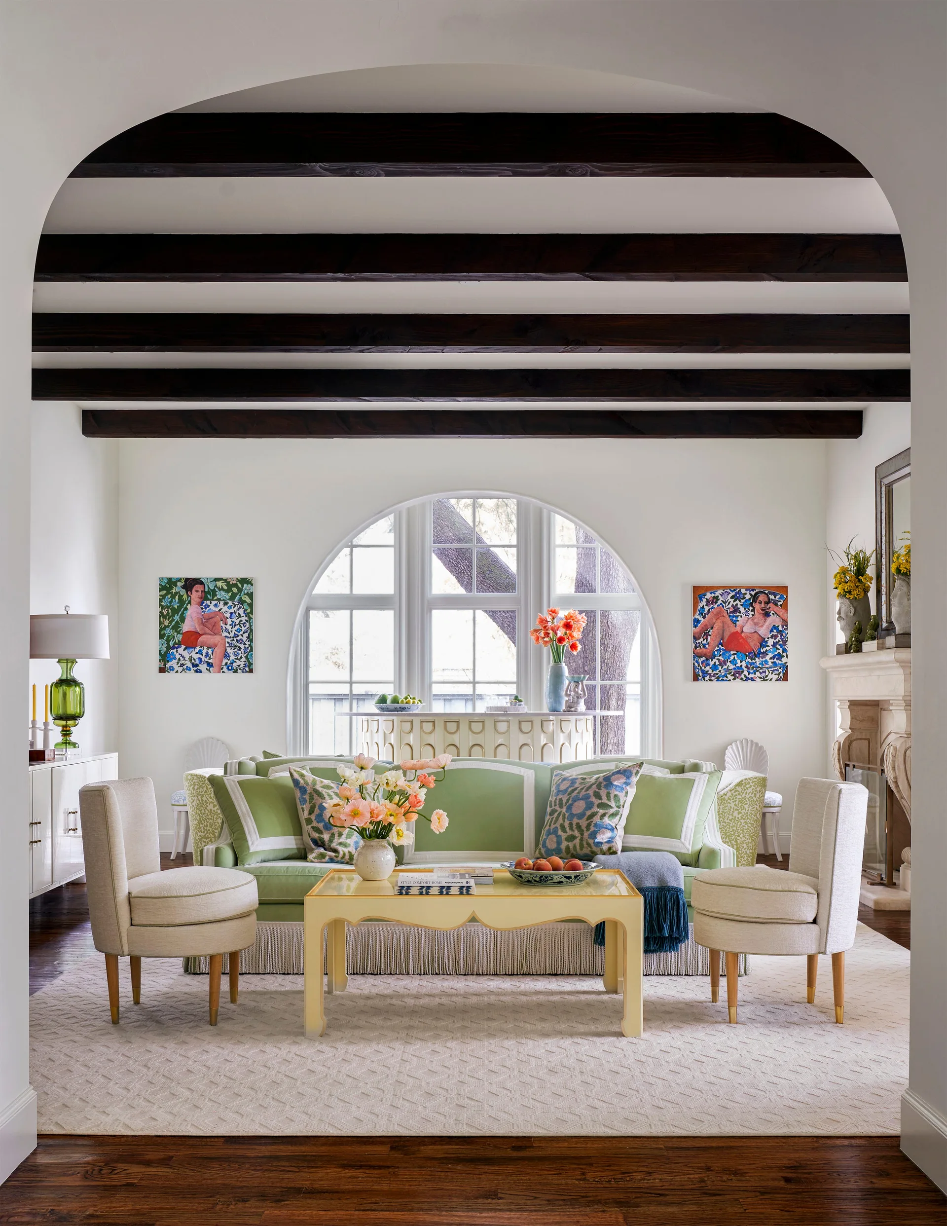 An airy living room with dark wooden ceiling beams, a green sofa, two cream armchairs, and a large arched window in the background.
