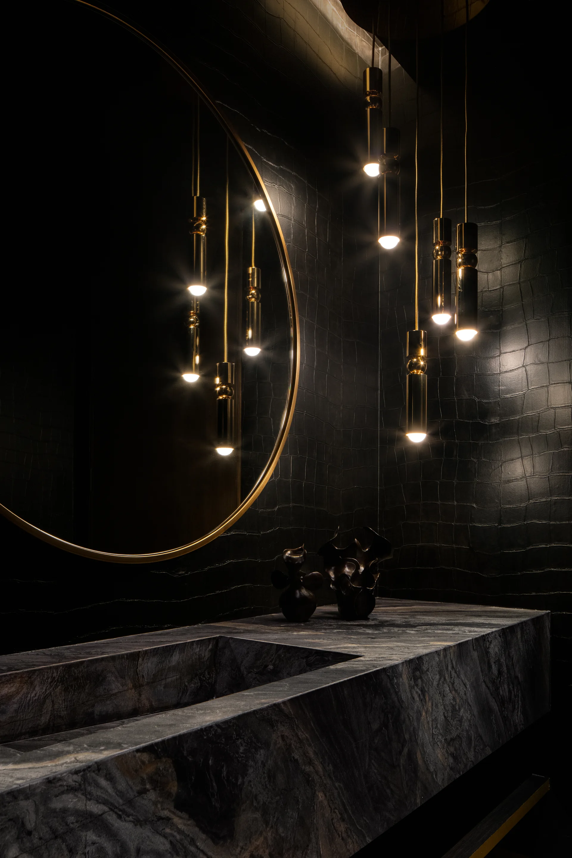 A dark powder room features a textured black wall, an integrated stone vanity, and a cluster of gold pendant lights reflected in a large circular mirror.