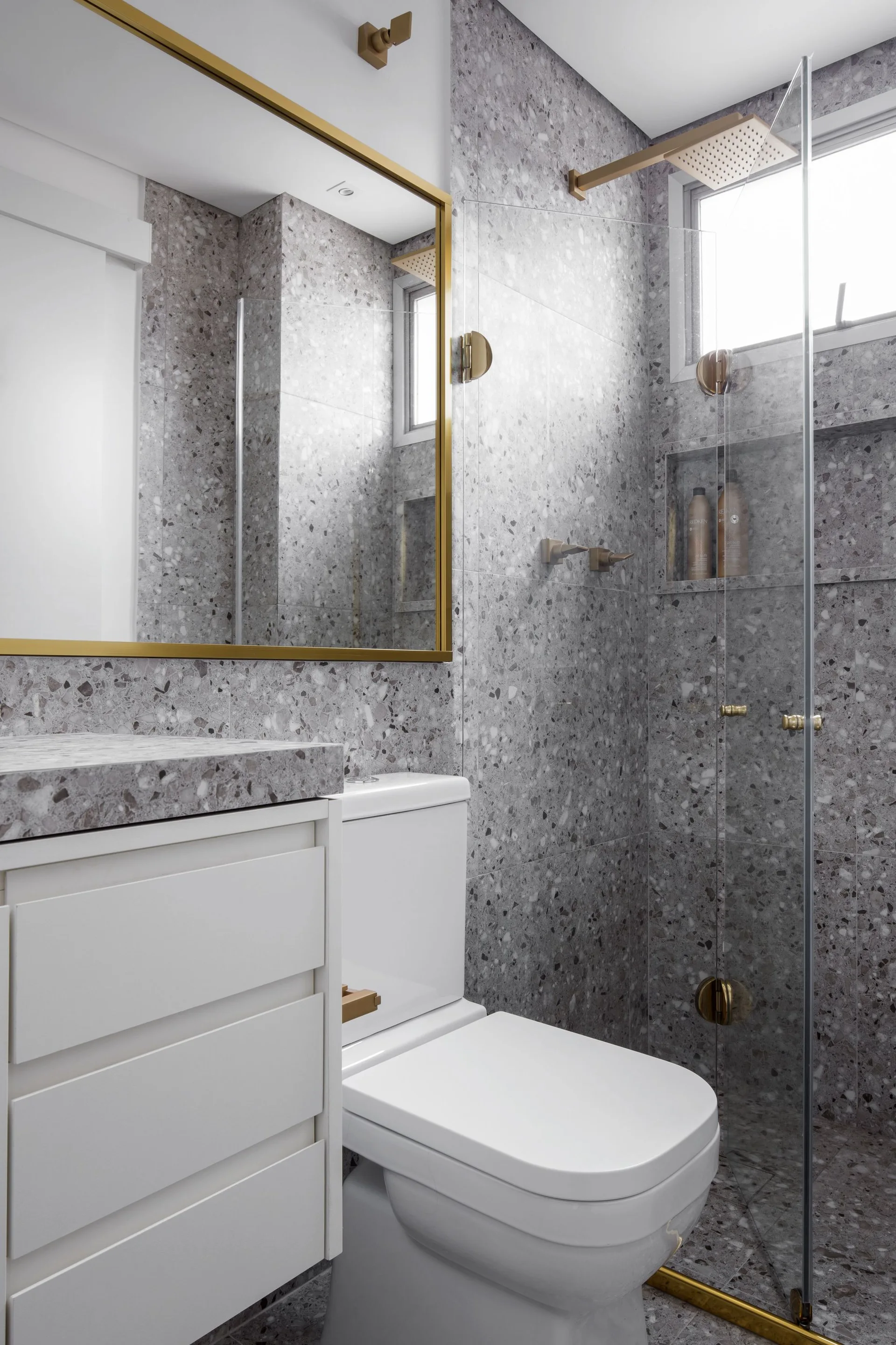 A modern bathroom features grey speckled terrazzo walls, a white vanity cabinet, a glass shower partition, and gold-trimmed fixtures.