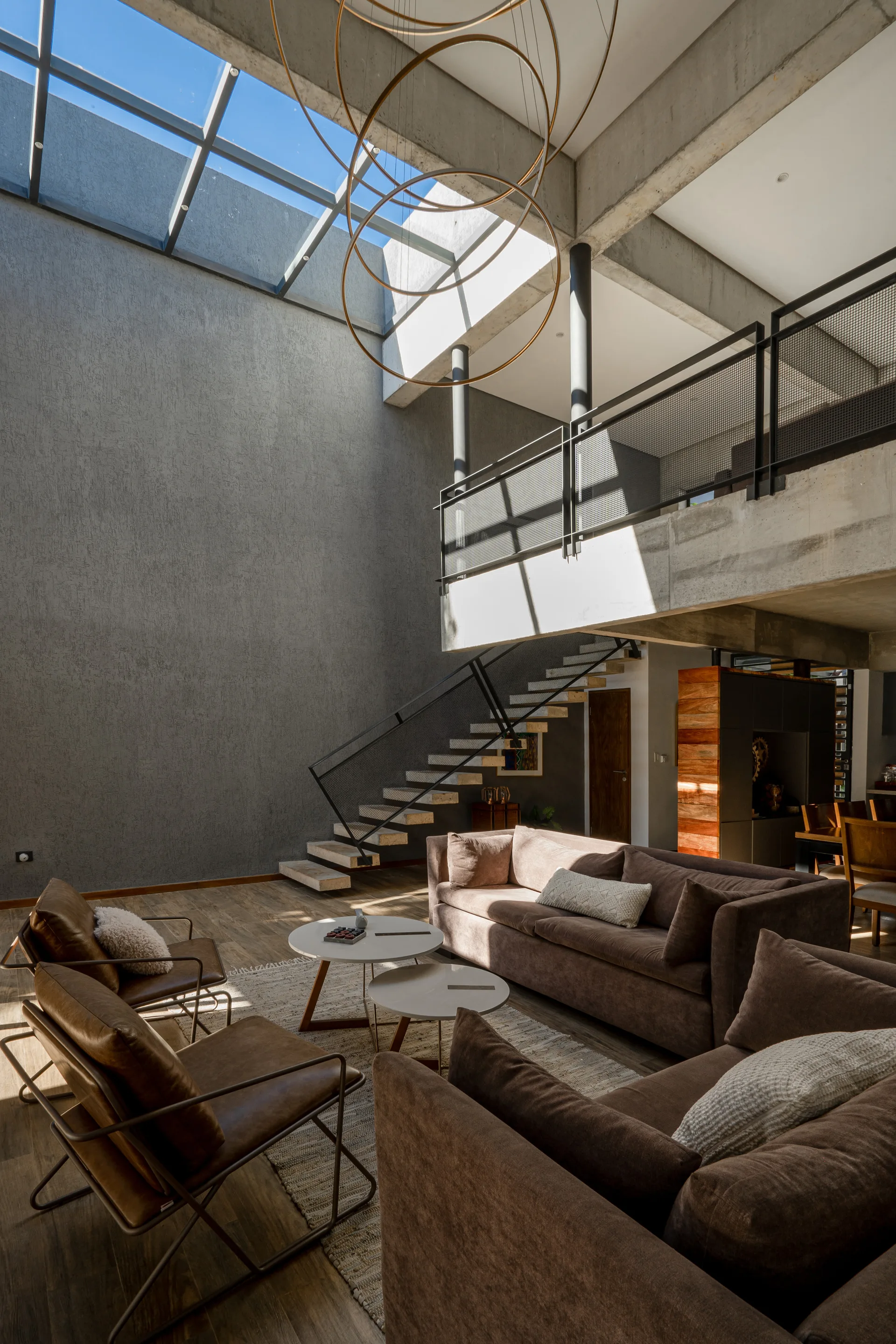 A double-height modern industrial living room features brown sofas, a metal-framed open staircase, concrete walls, and a large overhead skylight.