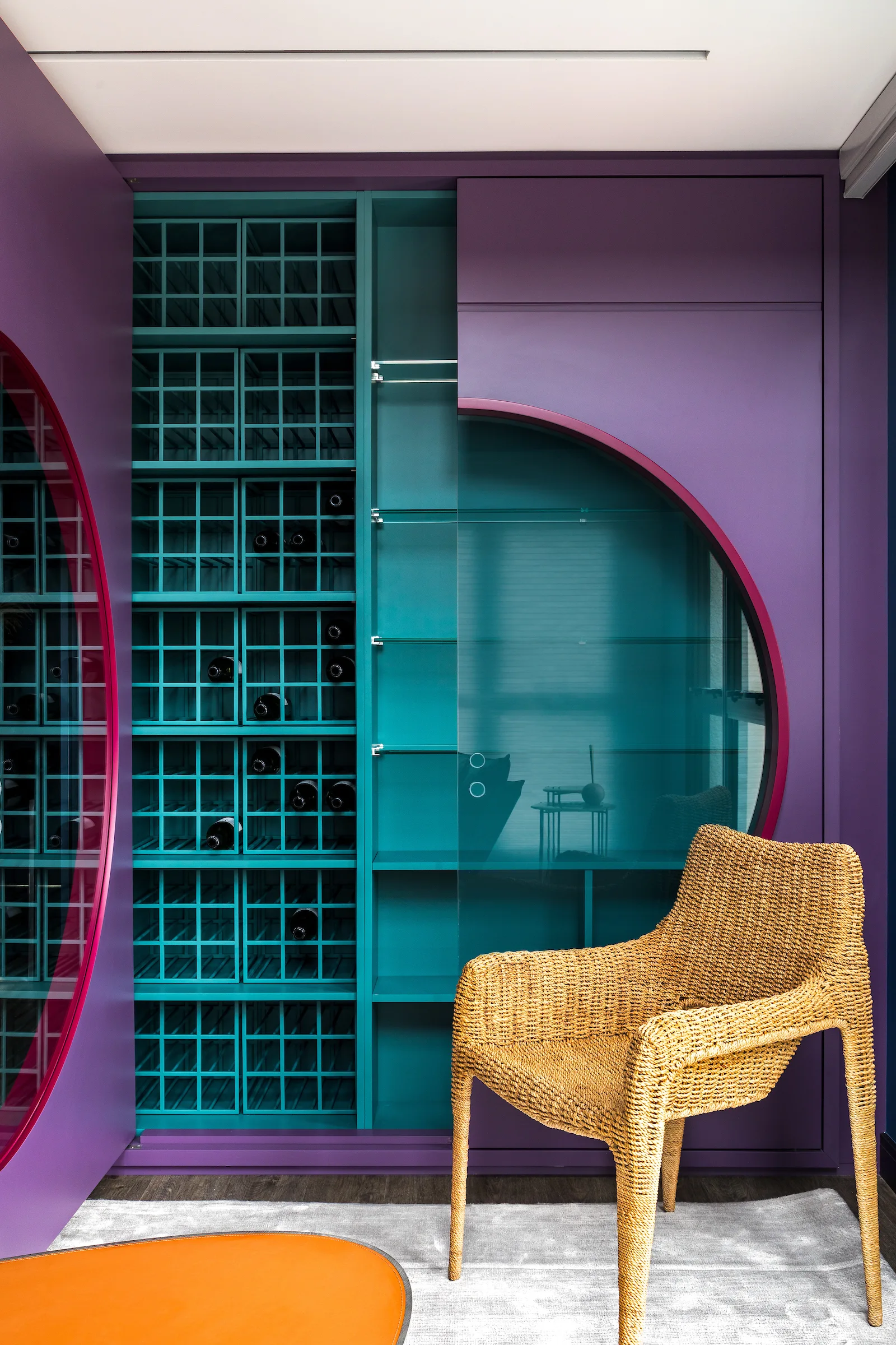 A bold purple and teal wine cabinet with a circular glass door stands behind a woven wicker armchair on a grey rug.