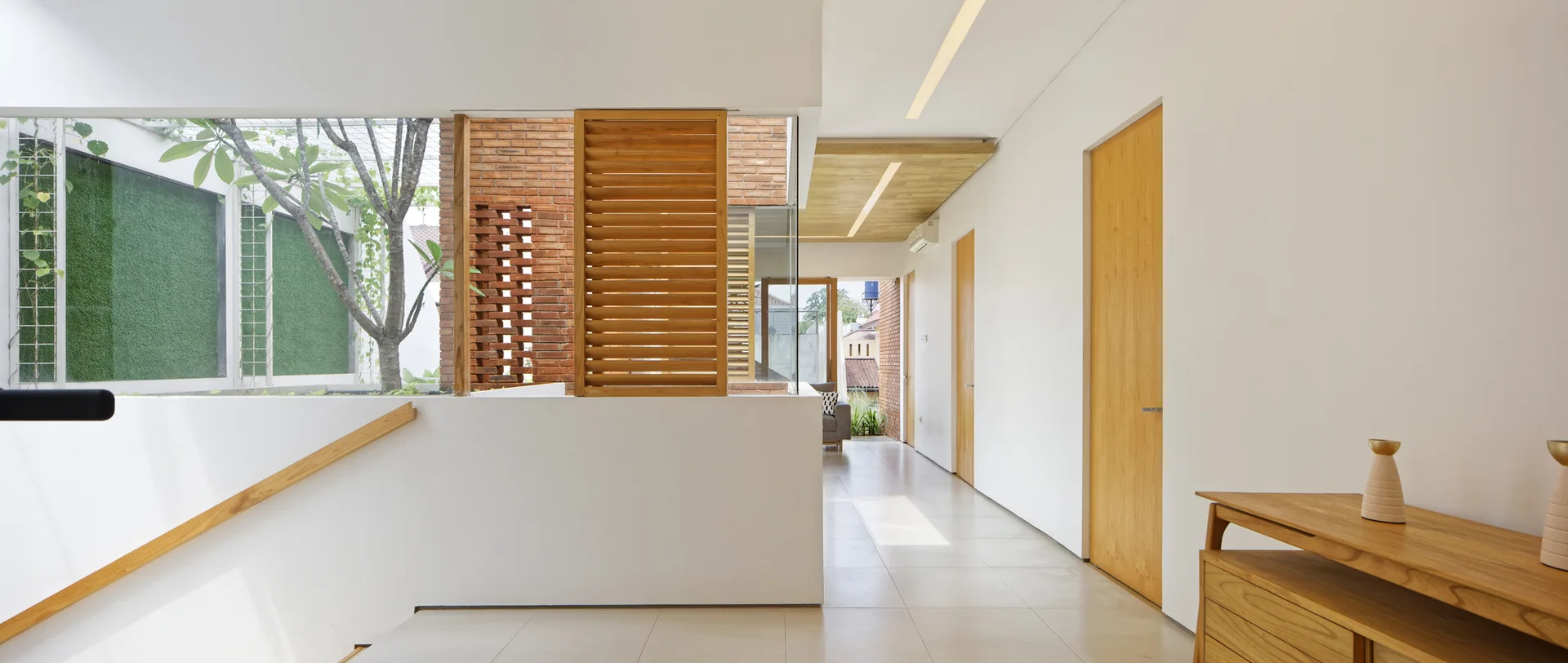 A bright modern hallway features smooth white walls, flush wood doors, an exposed brick partition with wooden shutters, and tiled flooring.