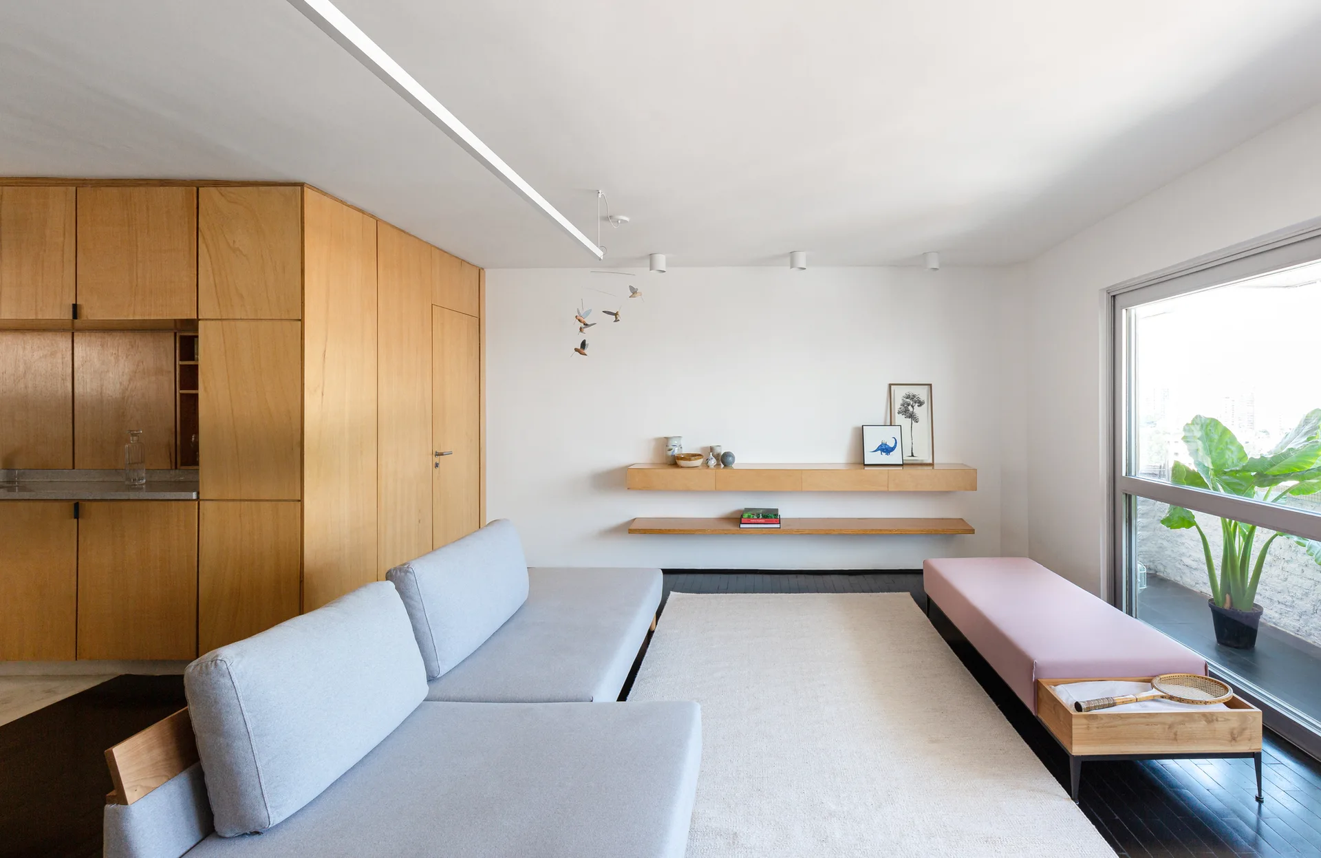 A minimalist living room features a grey sectional sofa, warm wood wall cabinetry, floating wooden shelves, and a pink bench on dark wood flooring.