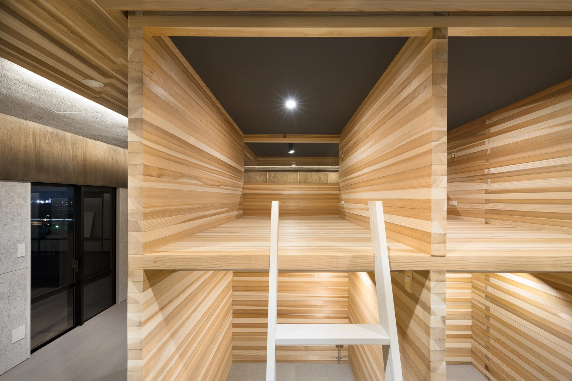 Built-in wooden sleeping pods feature horizontal wood slats on the walls, a dark ceiling with recessed spotlighting, and a white wooden ladder.