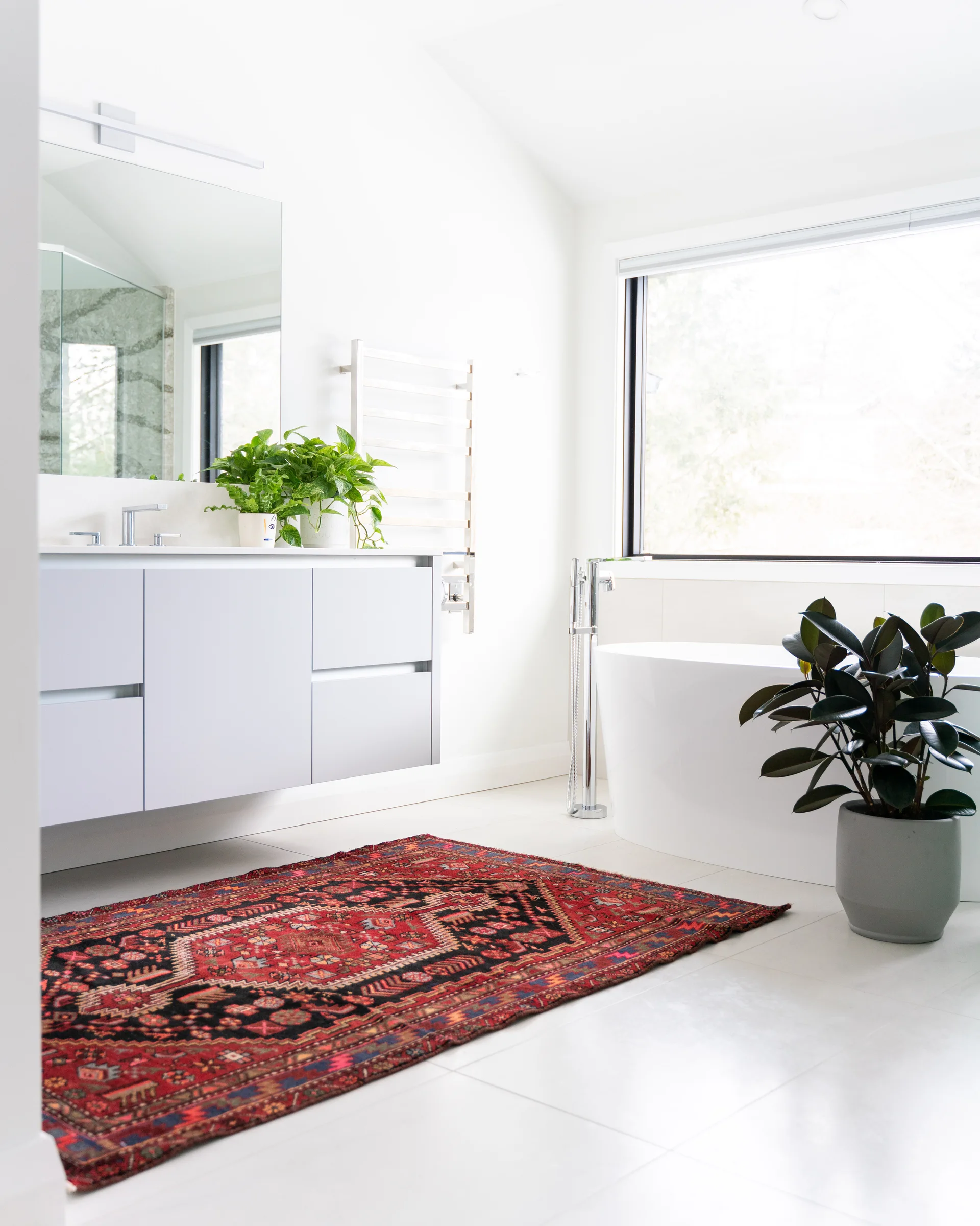 A bright white bathroom with a light grey floating vanity, a freestanding tub next to a large window, and an intricate red patterned area rug.