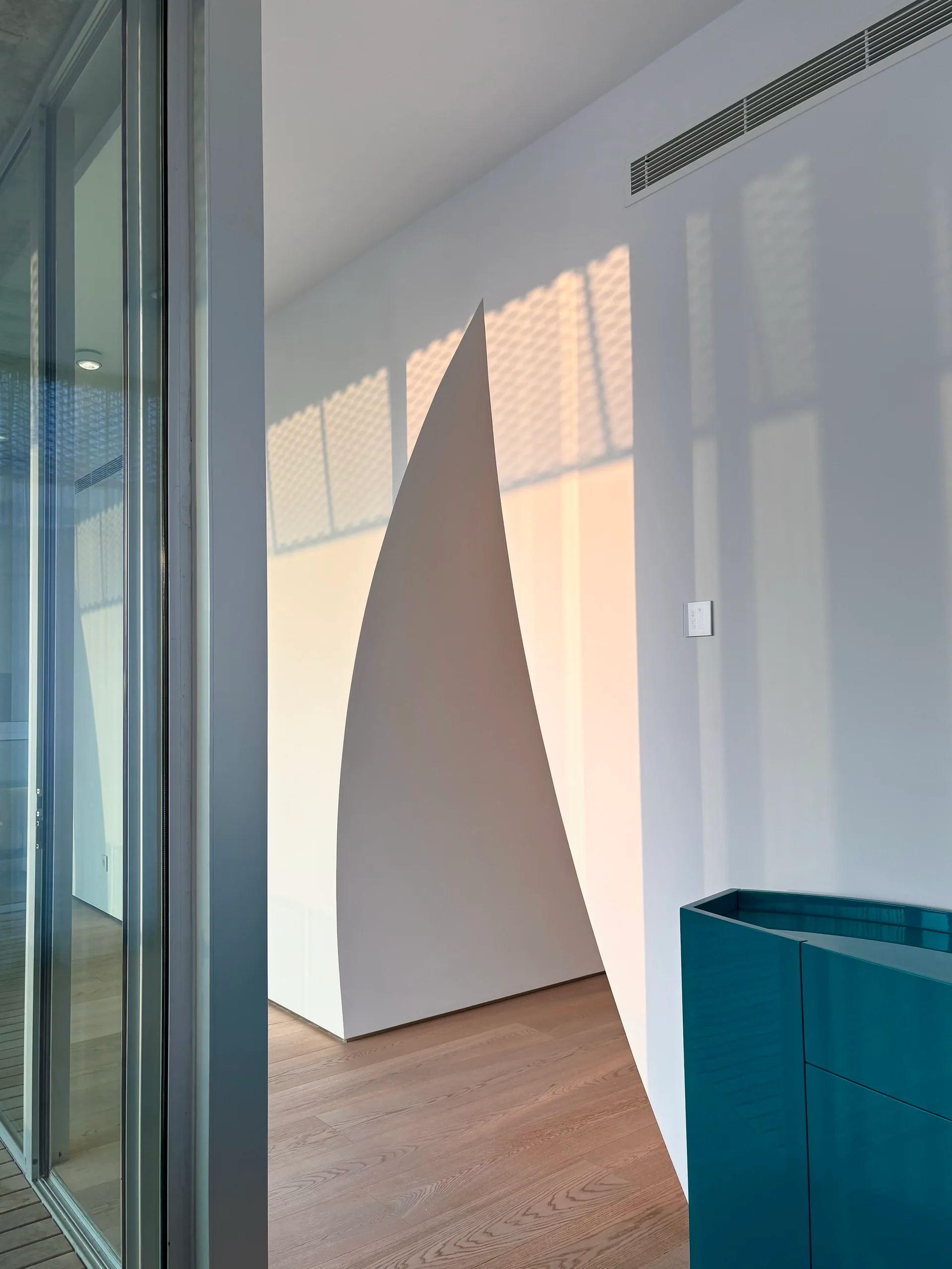 A minimalist interior featuring a white curved partition wall, a glossy teal cabinet on light wood floors, and warm sunlight.