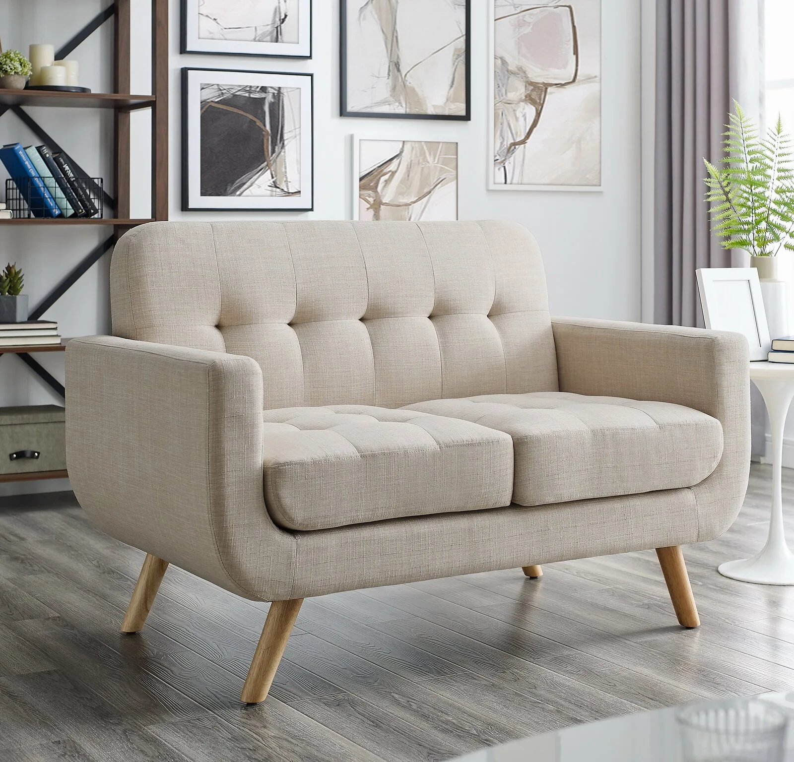 A beige tufted fabric loveseat with tapered wooden legs sits on grey wood flooring in a bright room.