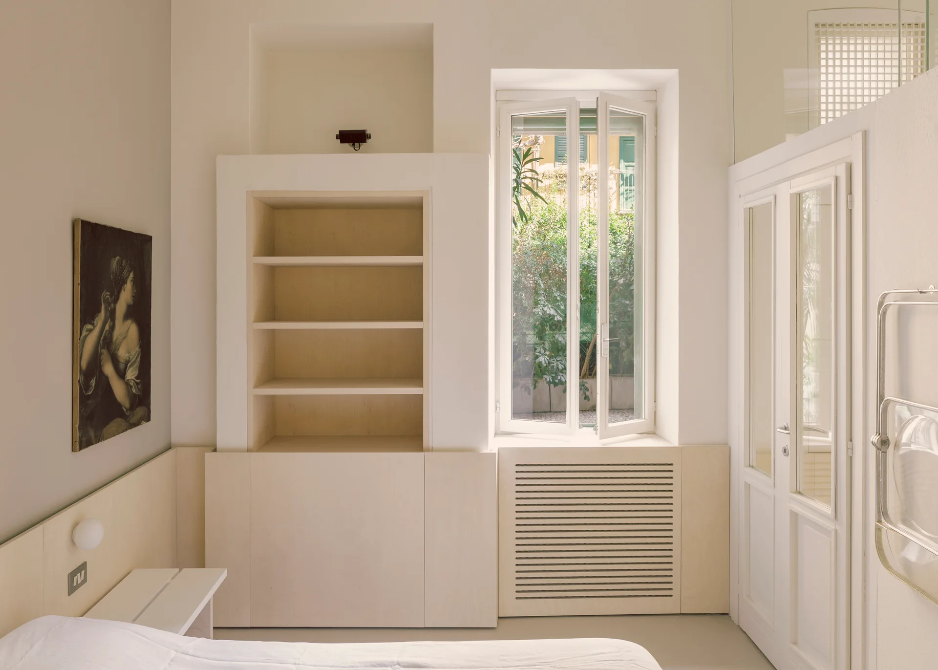 A minimalist bedroom featuring custom light wood built-in shelving, a slatted radiator cover beneath an open casement window, and low wall paneling.