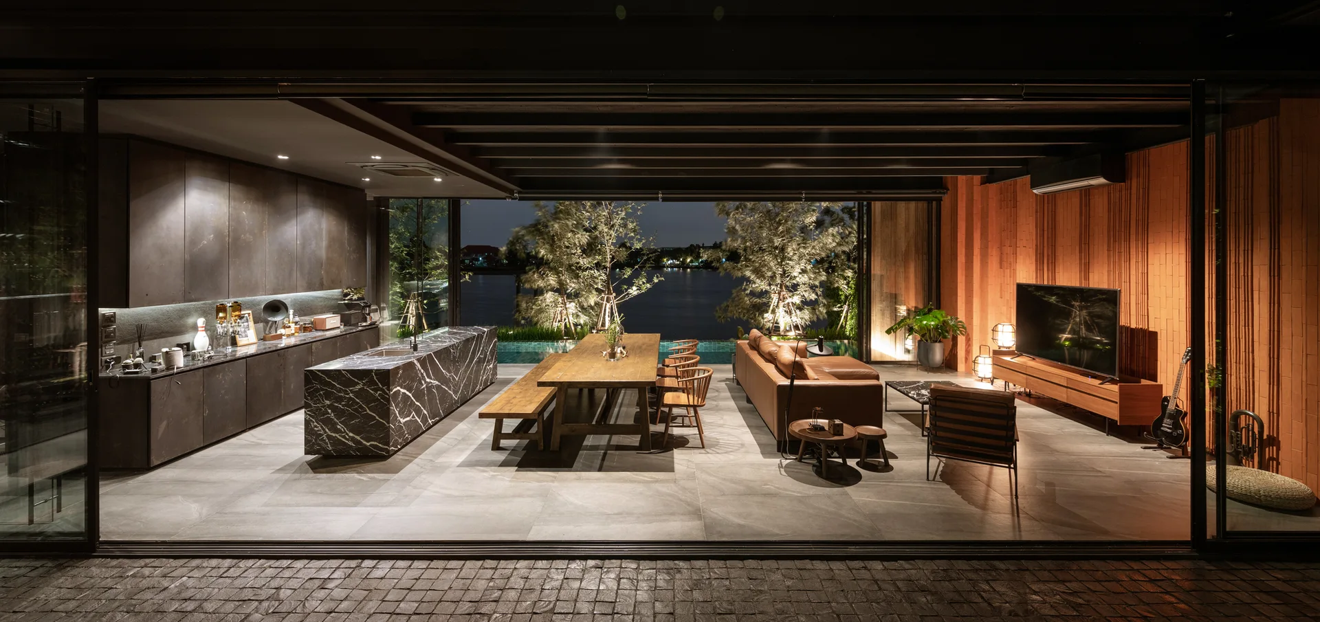 An open-concept living space features a black marble kitchen island, a wooden dining table, and a tan leather sofa facing a slatted wood wall.
