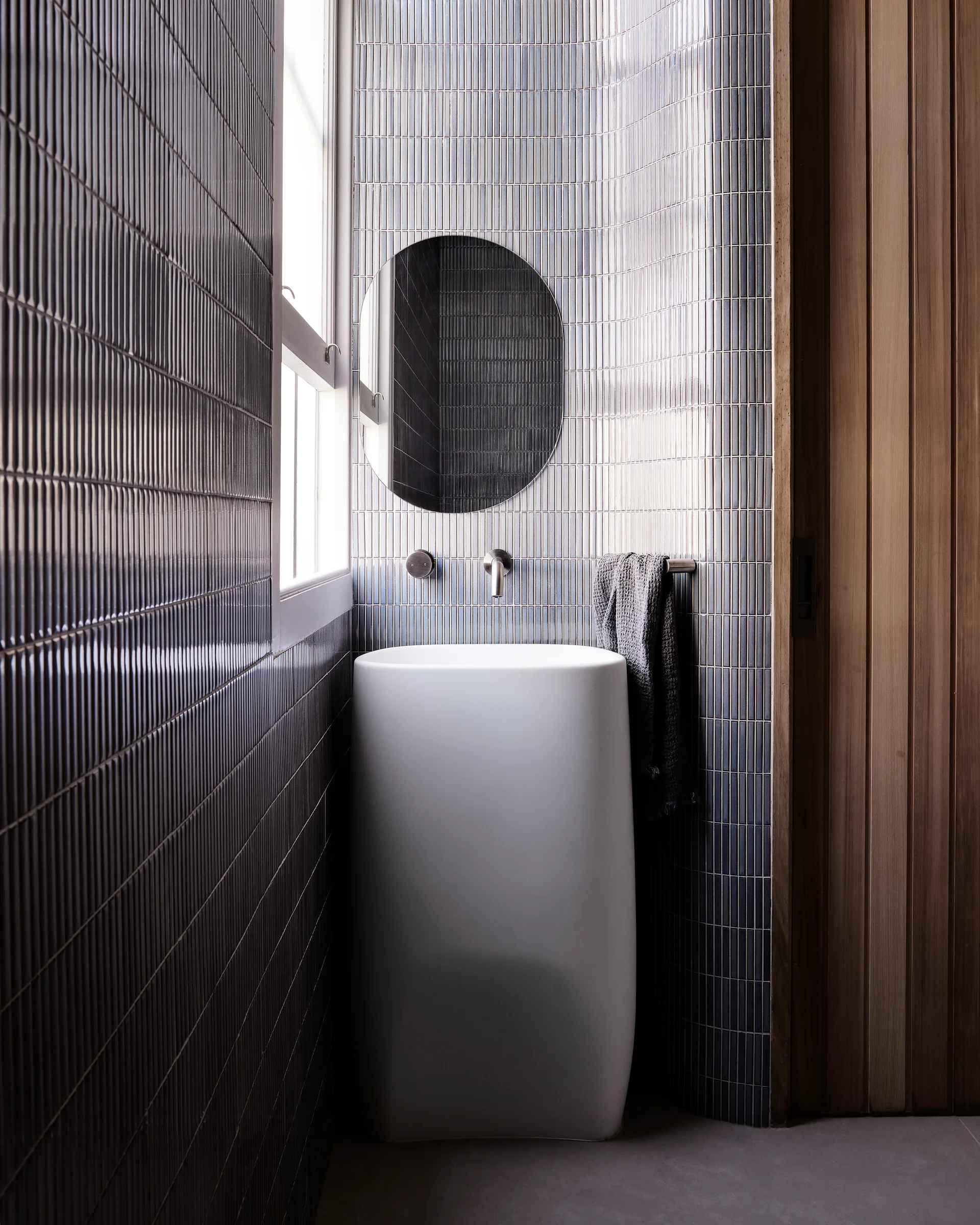 A compact powder room features a white monolithic pedestal sink set against curved walls lined with vertical blue-grey fluted tiles.