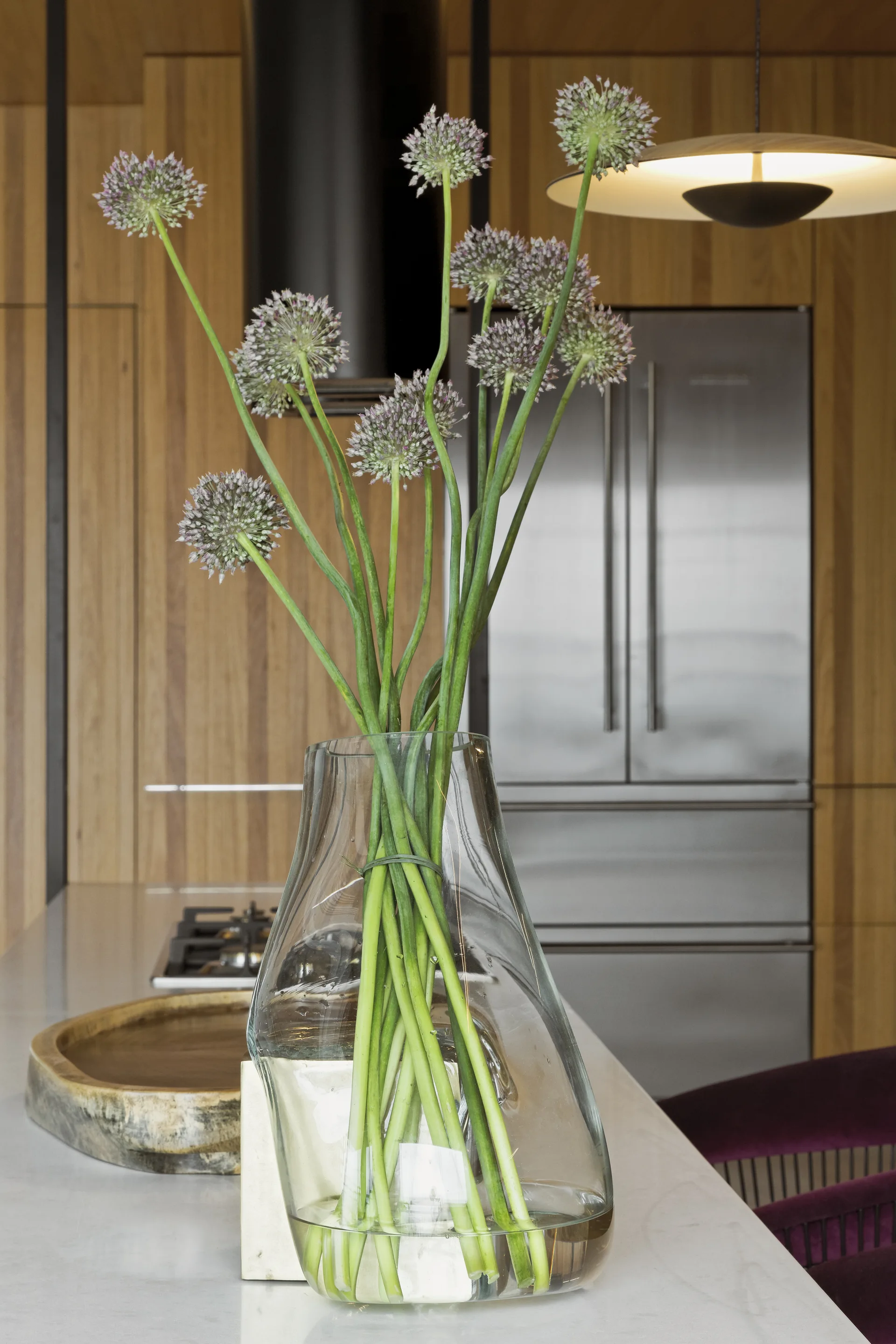 A clear glass vase holding purple allium flowers sits on a white marble kitchen island in front of vertical wood cabinetry.
