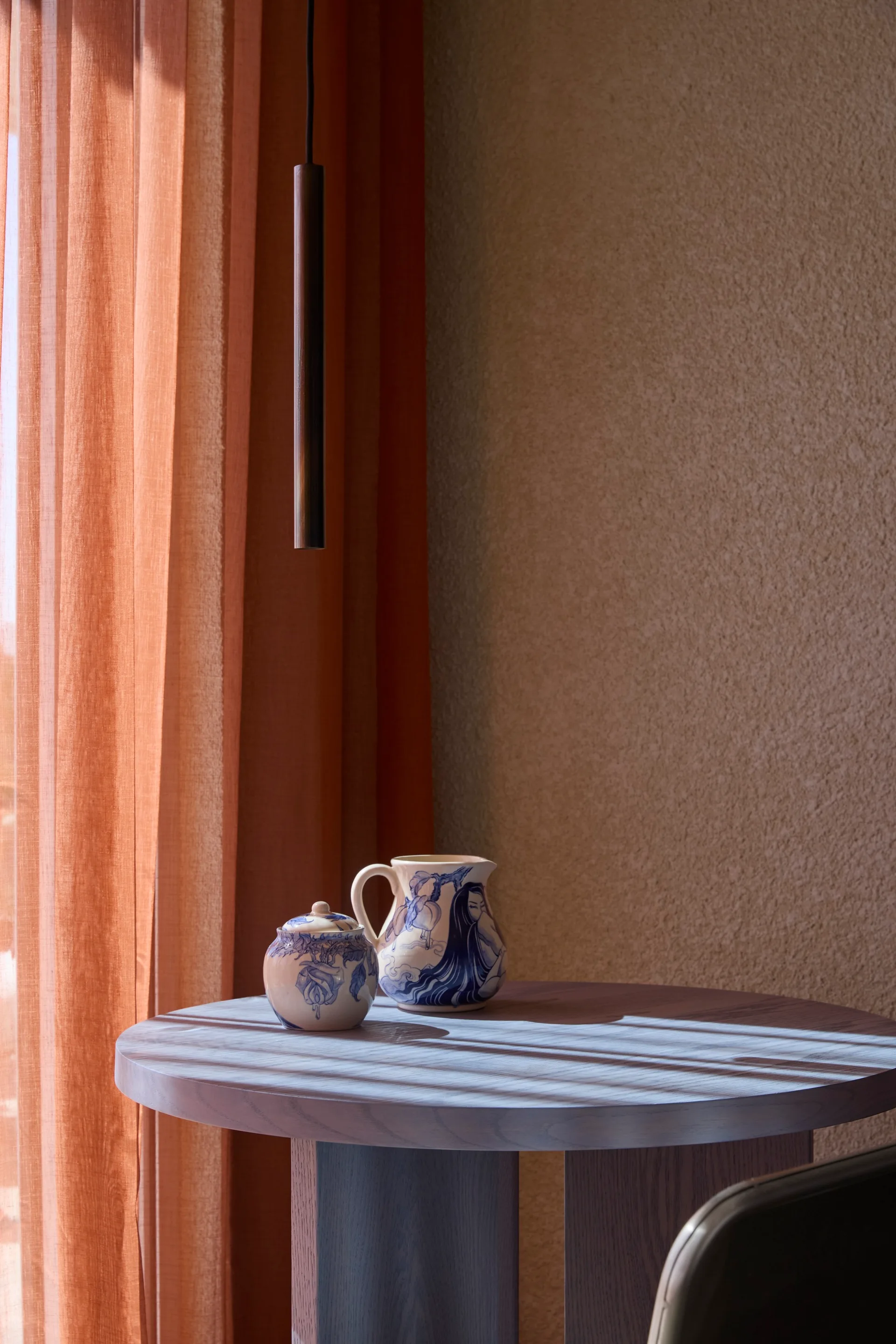 Blue hand-painted ceramics sit on a round wooden table cast with shadows from sheer terracotta curtains in front of a textured wall.