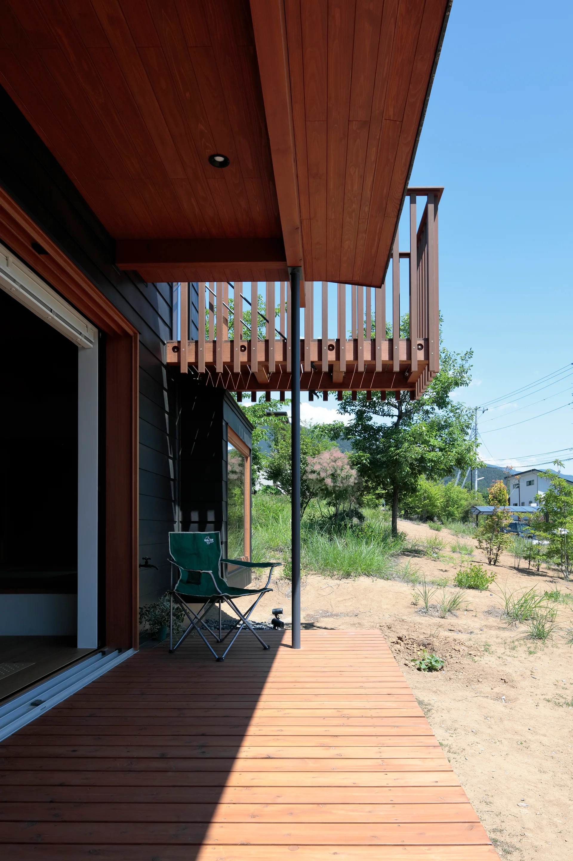 A modern wood deck beneath a matching planked ceiling overhang with a black support column and vertical wood balcony railing above.