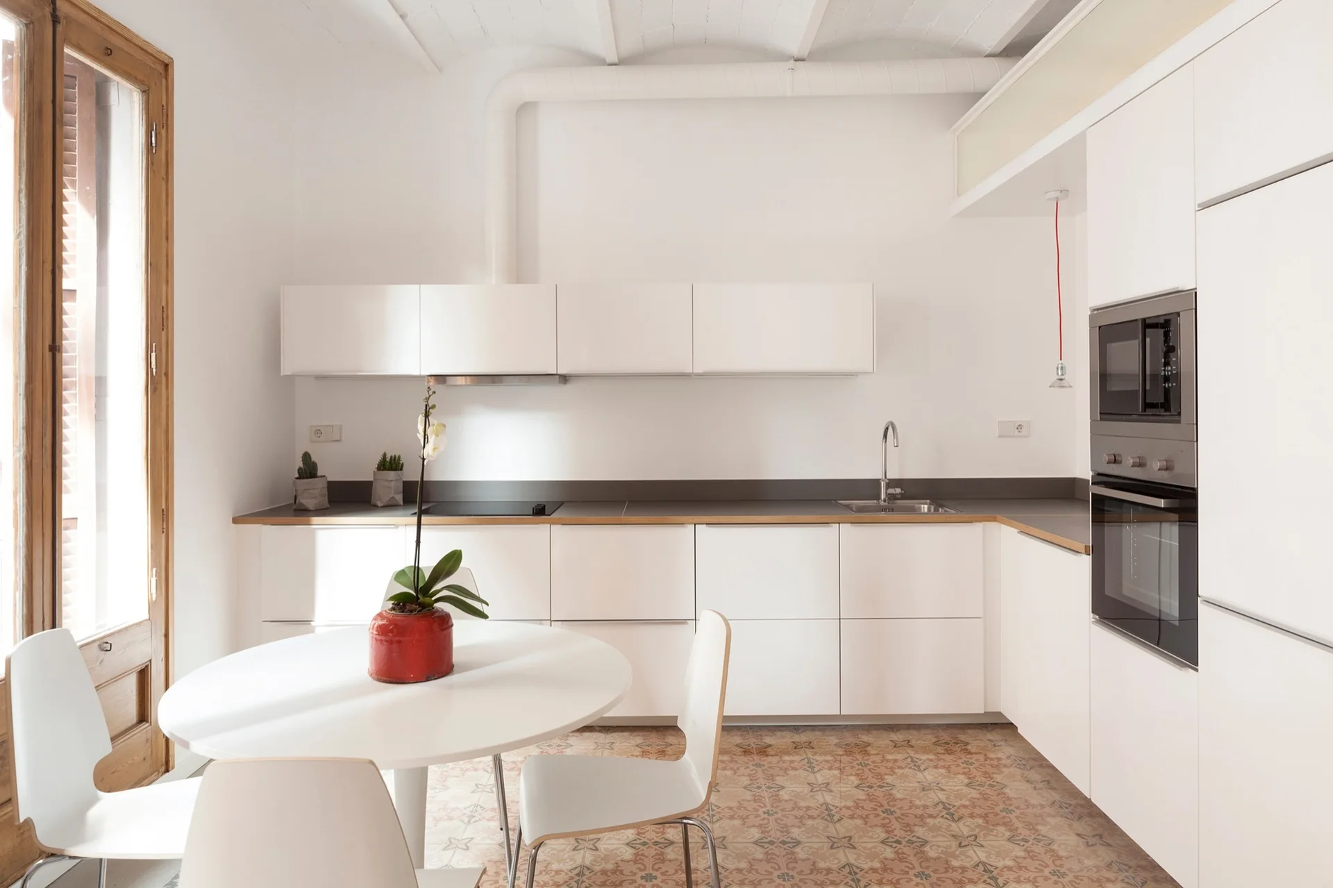 A minimalist white kitchen featuring flat-front cabinetry, a round dining table with chairs, and patterned terracotta floor tiles.