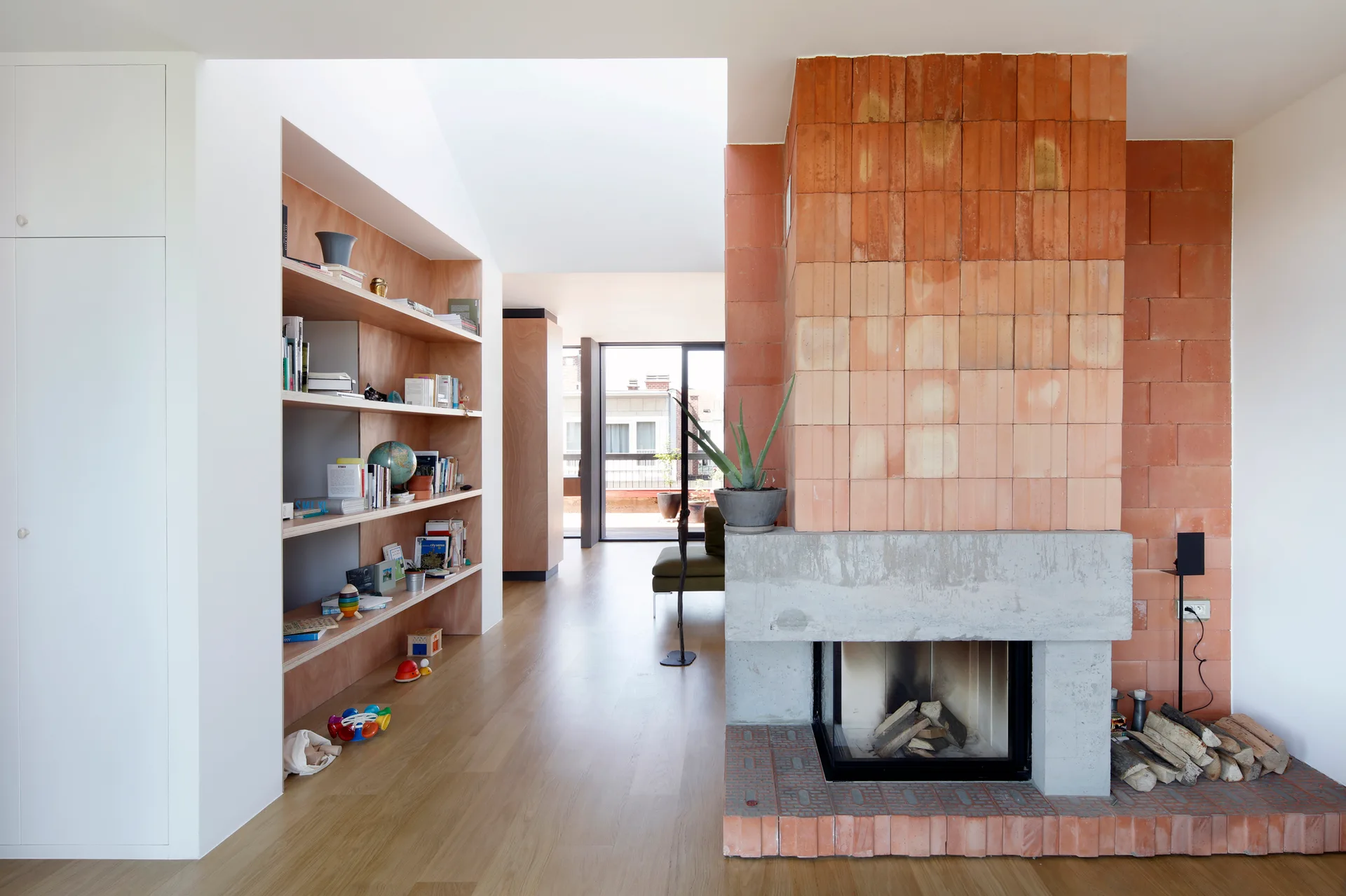 A modern living room features a tall fireplace clad in orange terracotta tiles with a concrete mantel, next to a built-in light wood bookshelf.