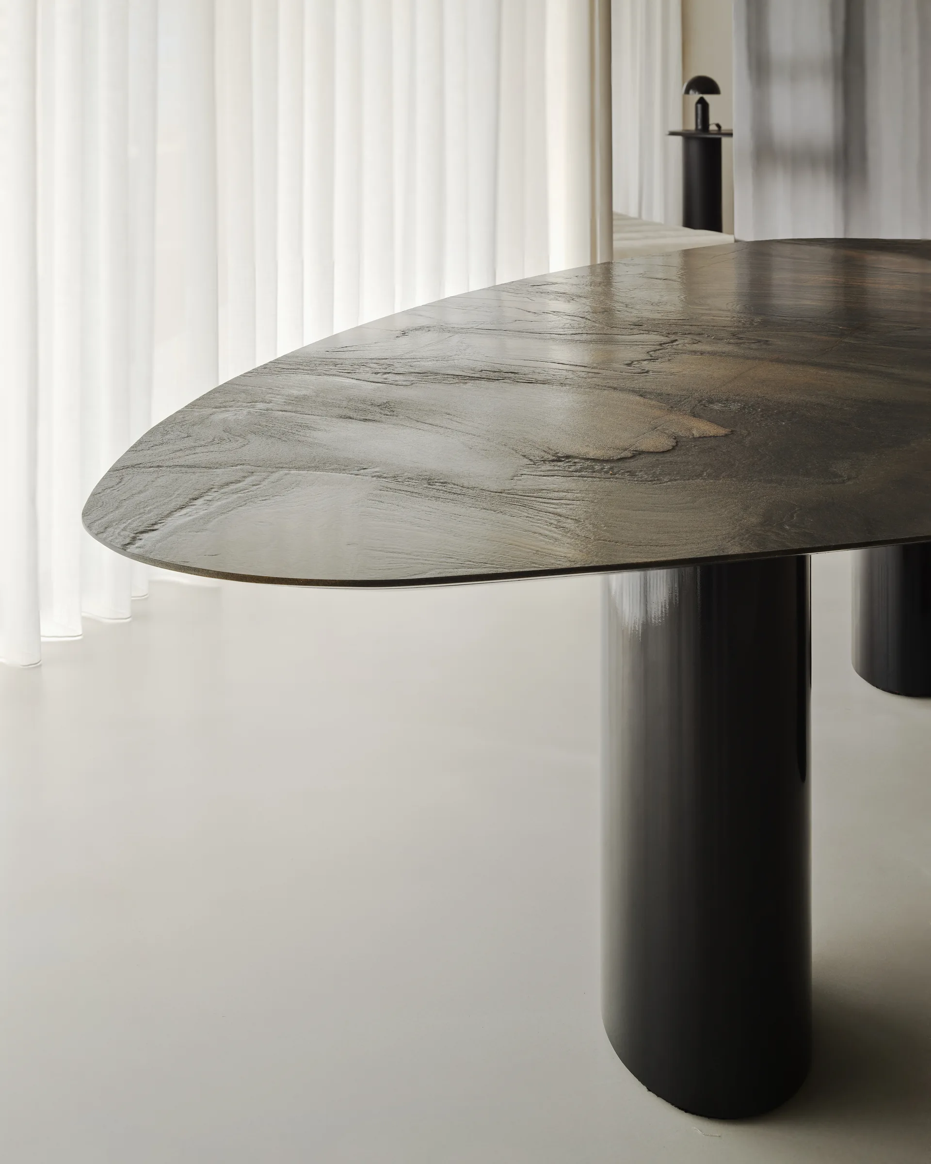 An organic-shaped dark dining table with a textured top and glossy black cylindrical legs stands in front of sheer white curtains.