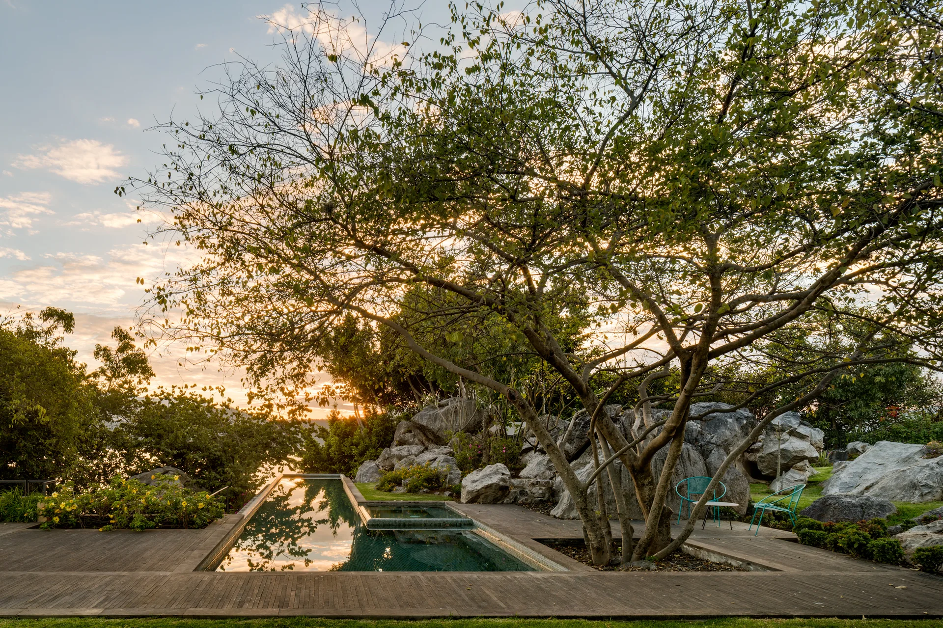 A rectangular swimming pool with dark tiles sits in a wood deck next to a large tree and massive natural rock formations at sunset.