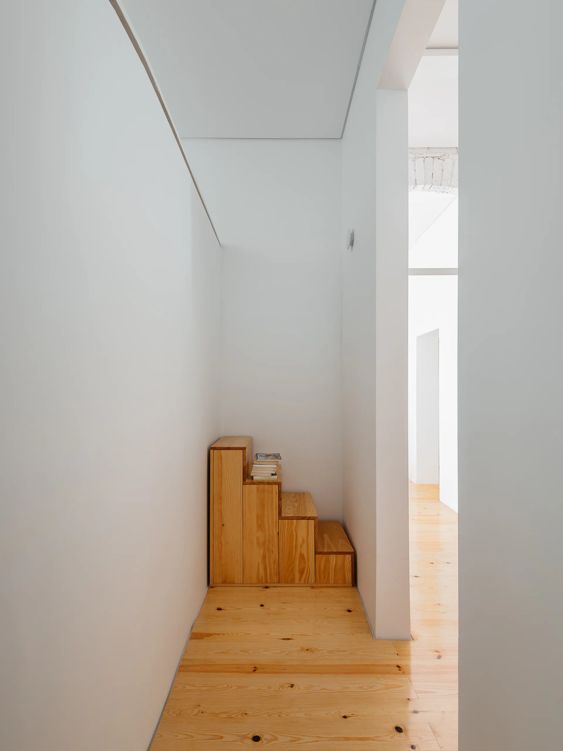 A narrow white hallway features natural pine wood floors and a small wooden stepped structure holding a stack of books against the wall.