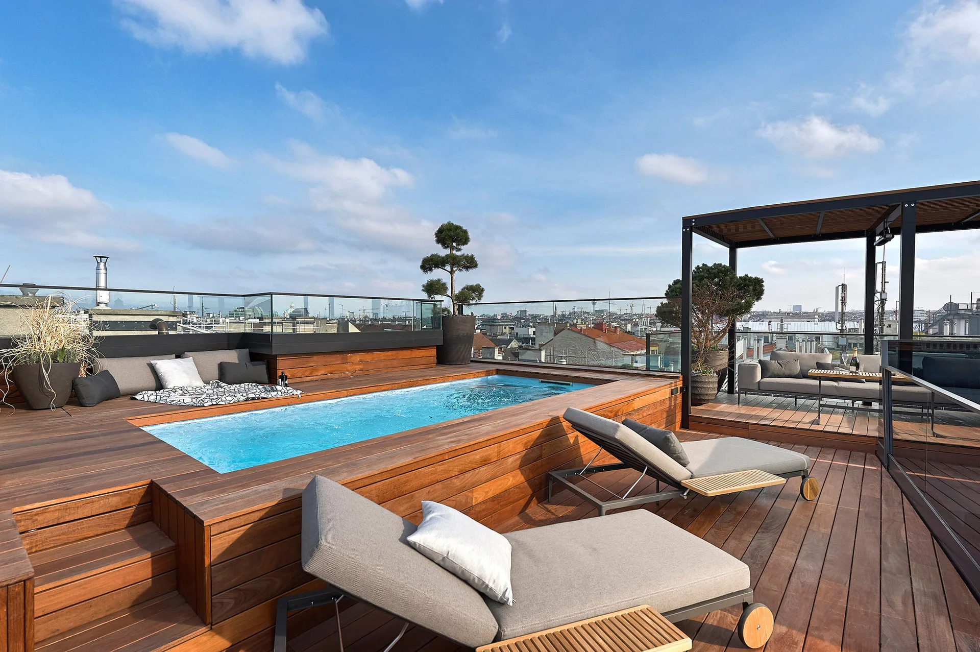 A wood-decked rooftop terrace features a rectangular swimming pool, grey chaise lounges, a glass railing, and a black metal pergola against a city skyline.