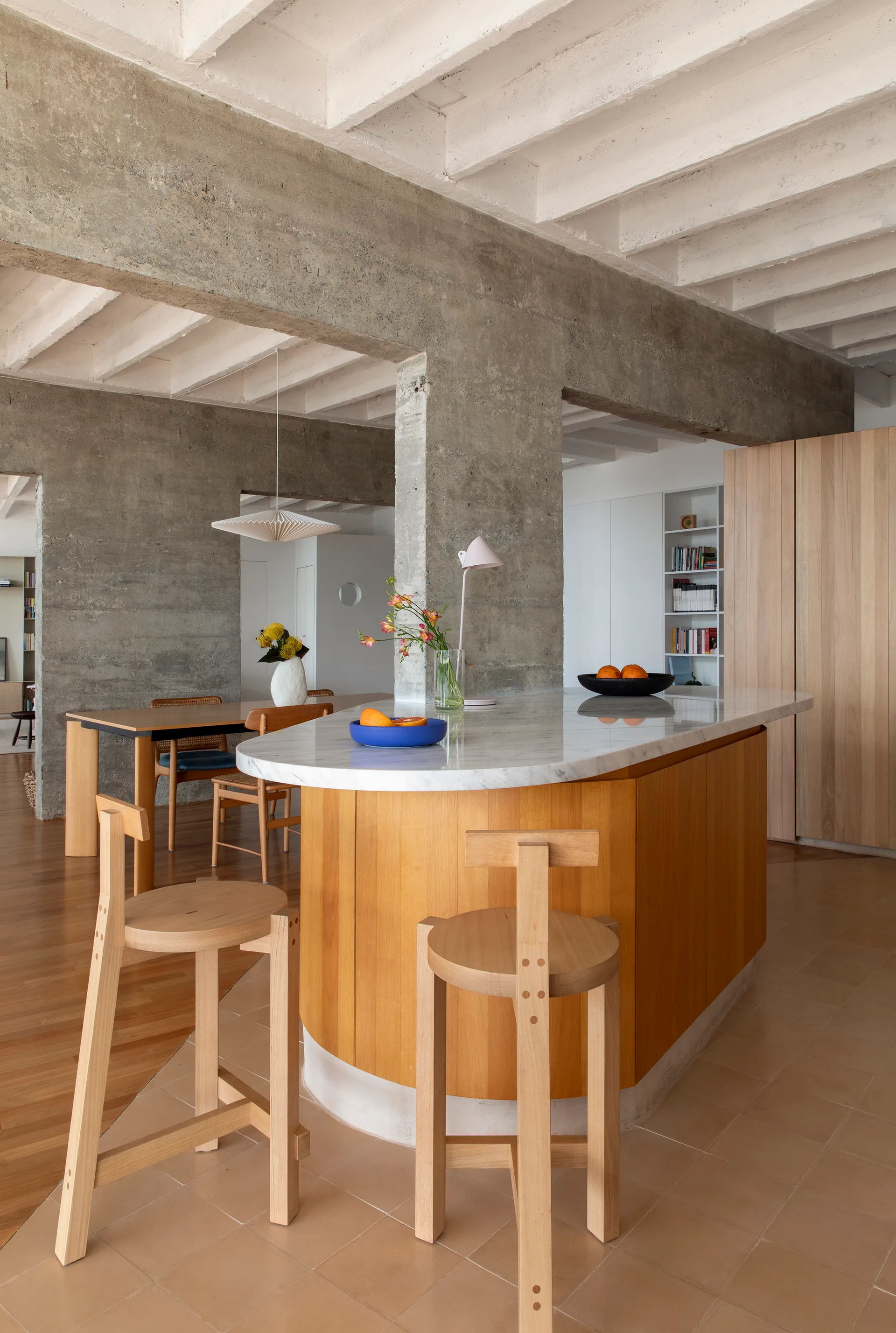 A curved wooden kitchen island with a marble countertop sits beneath exposed concrete beams alongside two minimal wood bar stools.