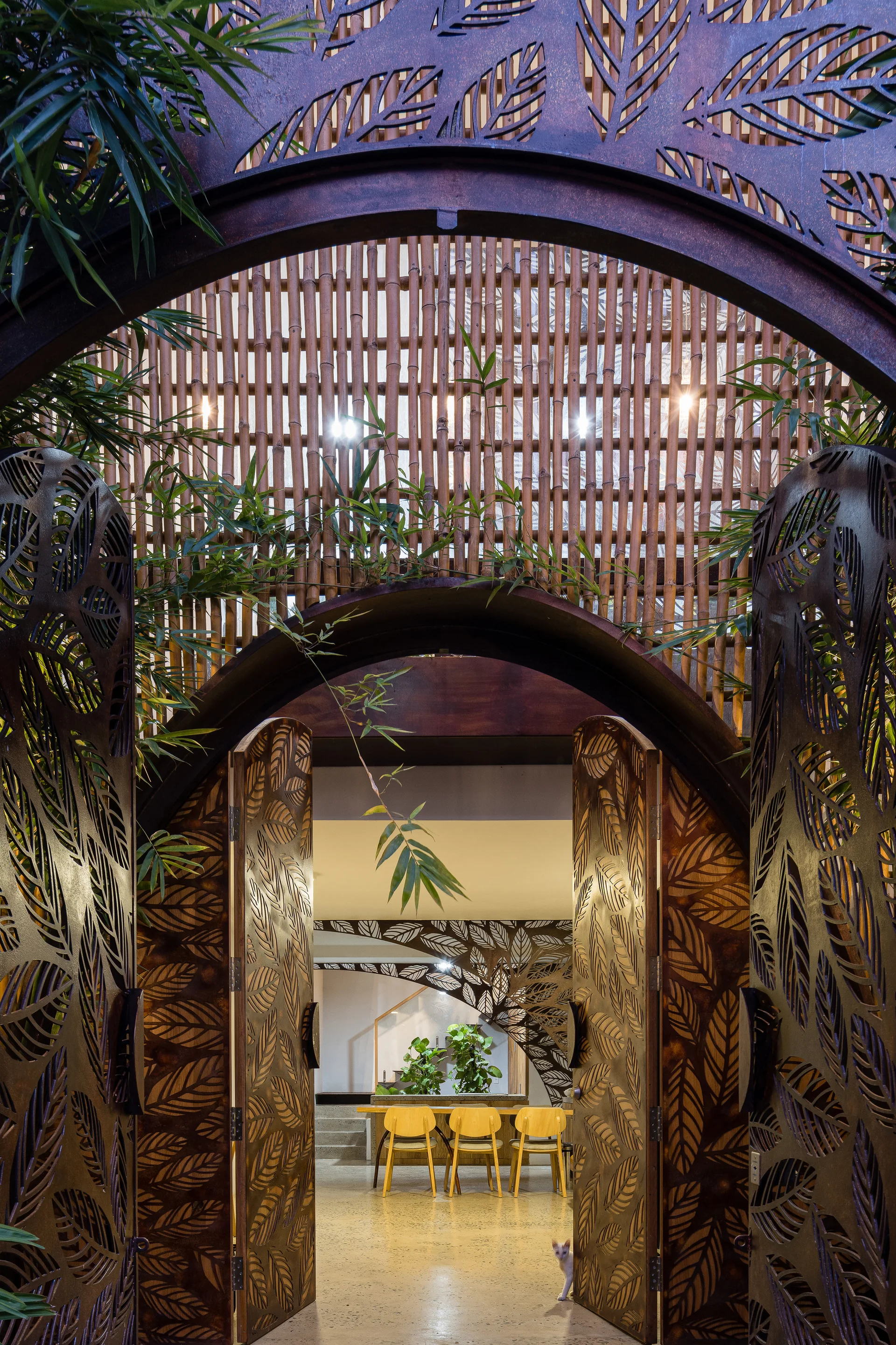 An arched entryway with ornate leaf-patterned metal gates opens to reveal a dining area with wood chairs and a vertical bamboo screen backdrop.