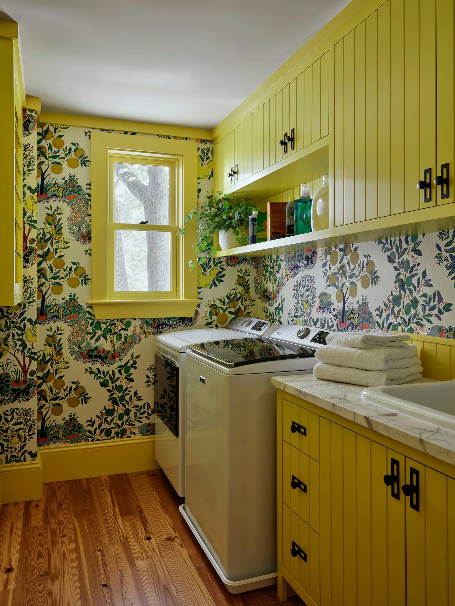 A laundry room featuring bright yellow tongue-and-groove cabinetry, botanical wallpaper, white washer and dryer, and pine wood flooring.
