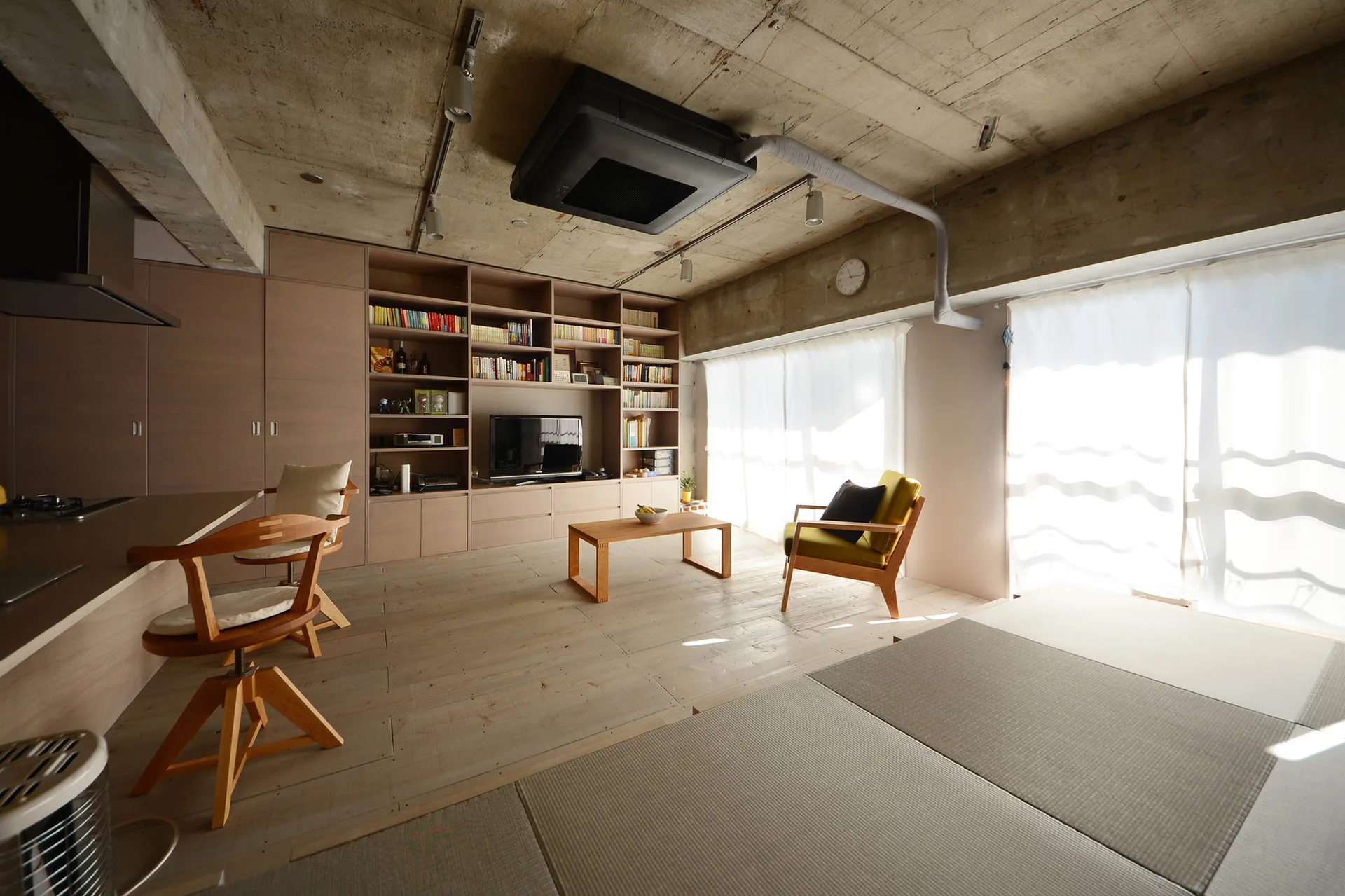 An open living room features an exposed concrete ceiling, a large light-wood bookshelf wall, and a raised floor section covered in tatami mats.