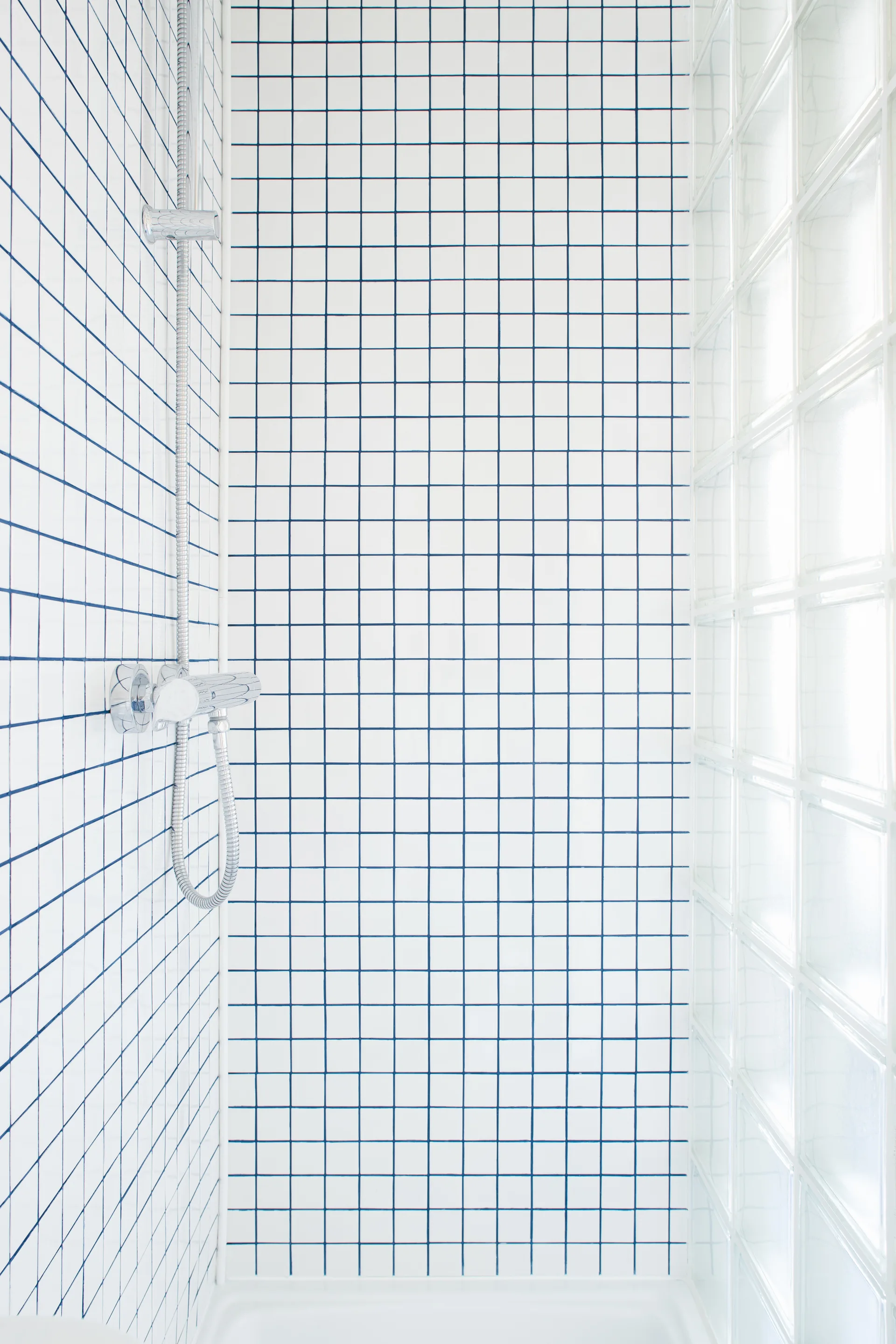 A shower interior features white square tiles with blue grout on the walls and a translucent glass block partition on the side.