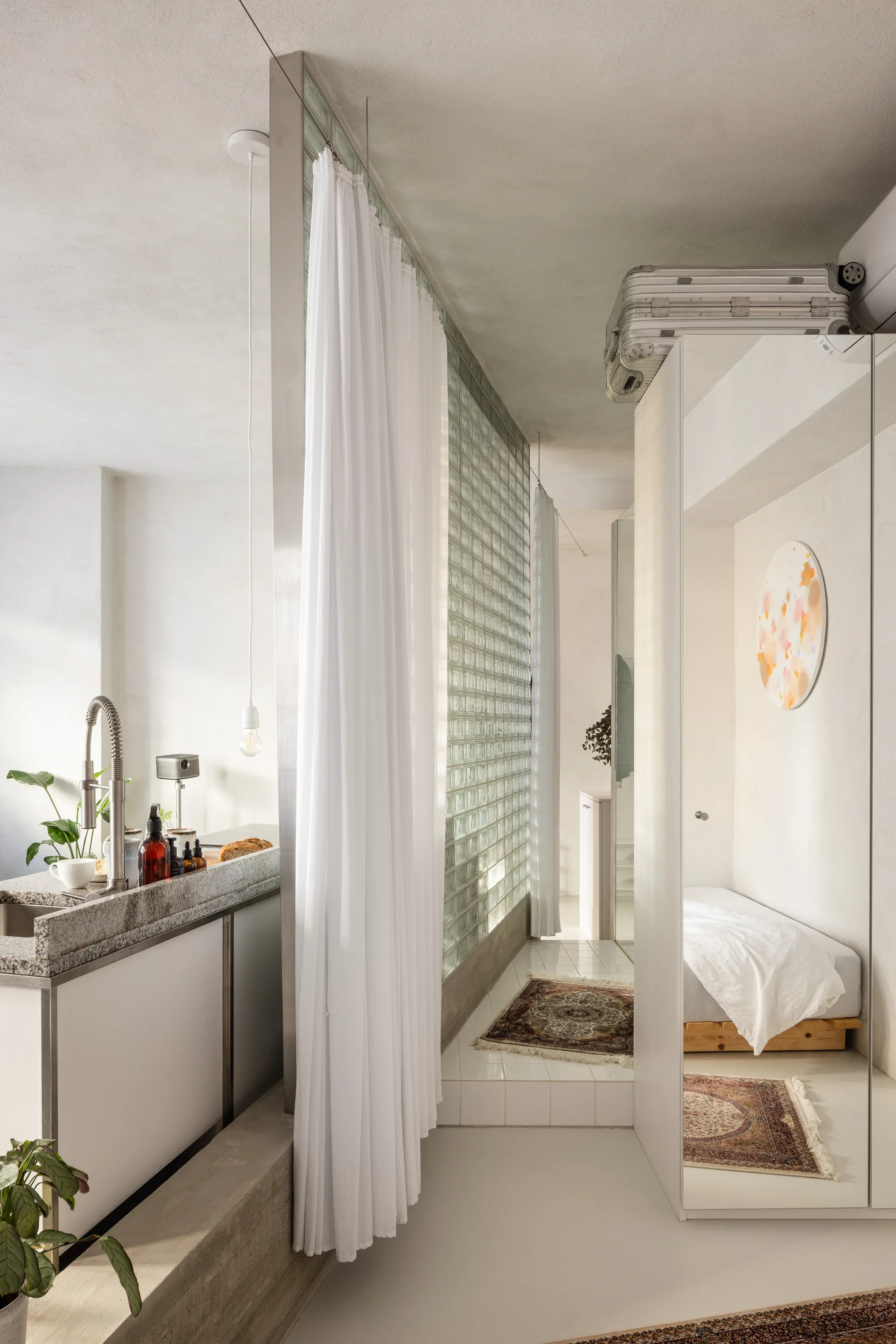 A minimalist studio interior features a kitchen island separated from a small bedroom area by a glass block wall and white curtains.
