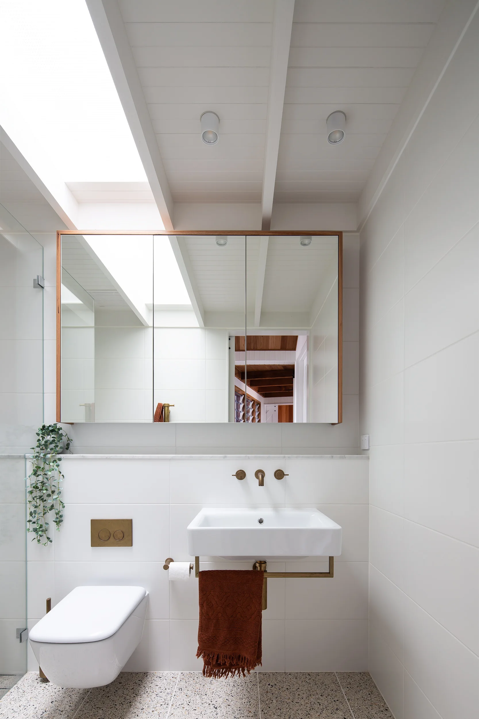 A modern white bathroom features a wall-mounted sink, a wood-framed mirror under a skylight, and speckled terrazzo tile flooring.