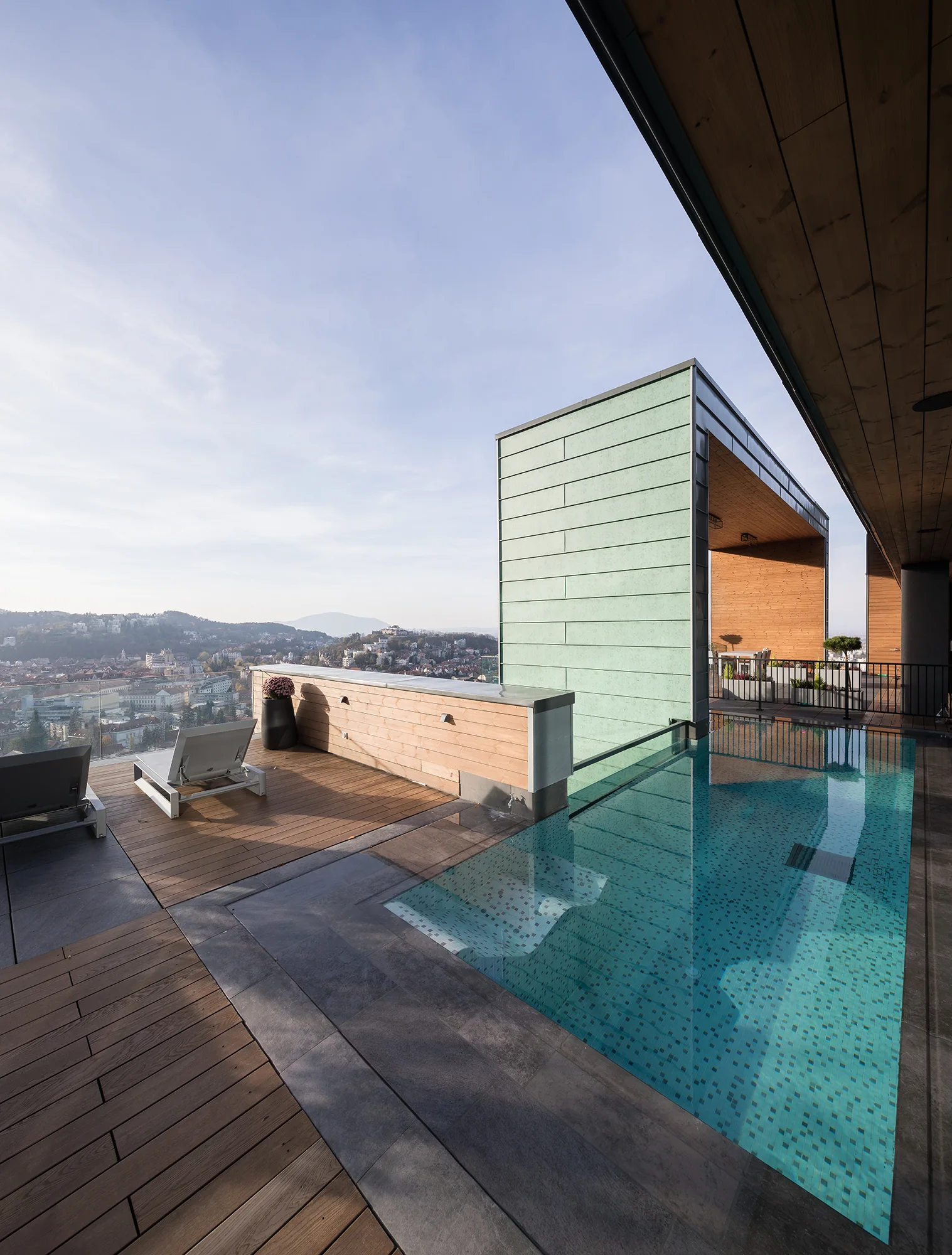An outdoor rooftop swimming pool features wooden decking, lounge chairs, and a large green-paneled structure overlooking a city.