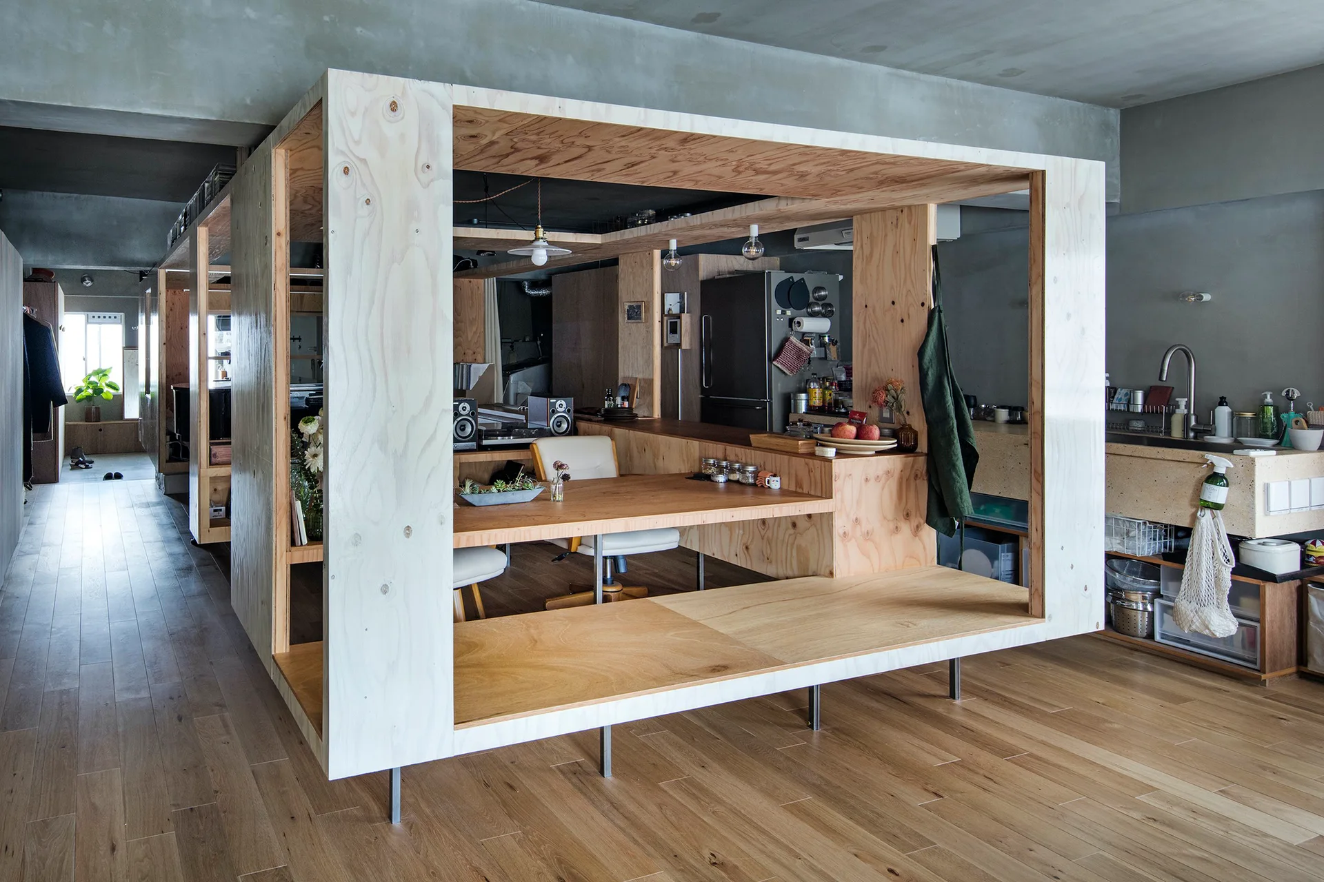 A freestanding plywood box structure with an integrated desk stands on wooden floors inside a gray loft apartment with concrete ceilings.