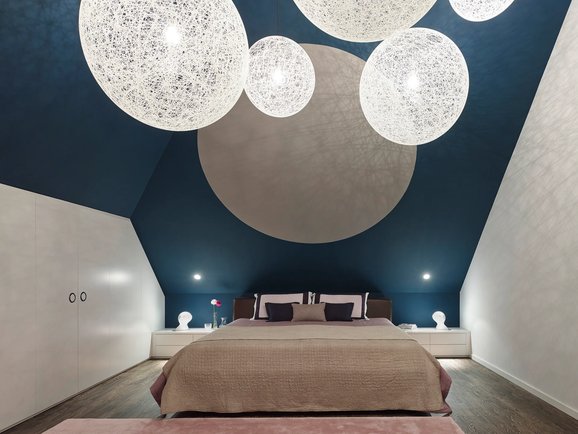 A modern attic bedroom with a low-profile bed beneath a dark blue sloped ceiling decorated with large white spherical pendant lights.
