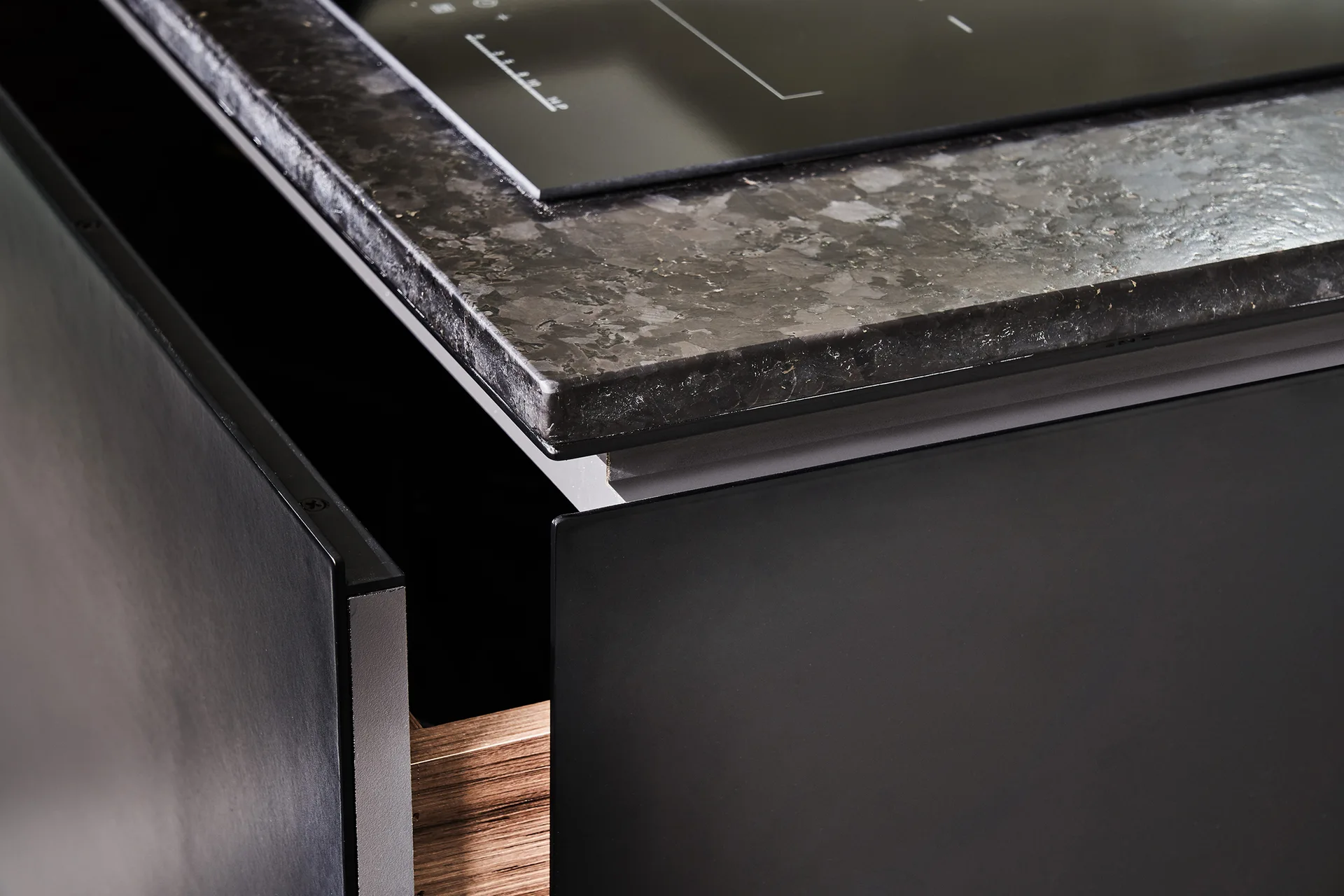 A close-up of a matte black kitchen cabinet with an open wooden drawer below a mottled dark grey stone countertop and black induction cooktop.