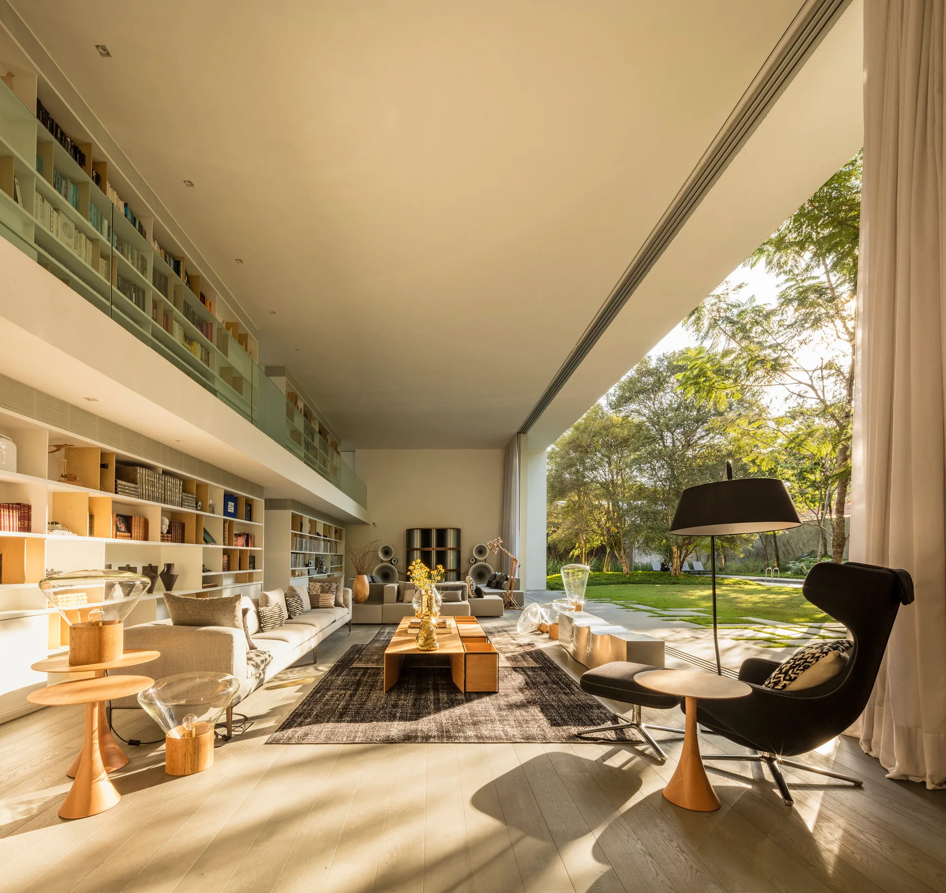 A double-height modern living room features a massive built-in bookcase wall, a low wooden coffee table, and an open wall facing a green lawn.