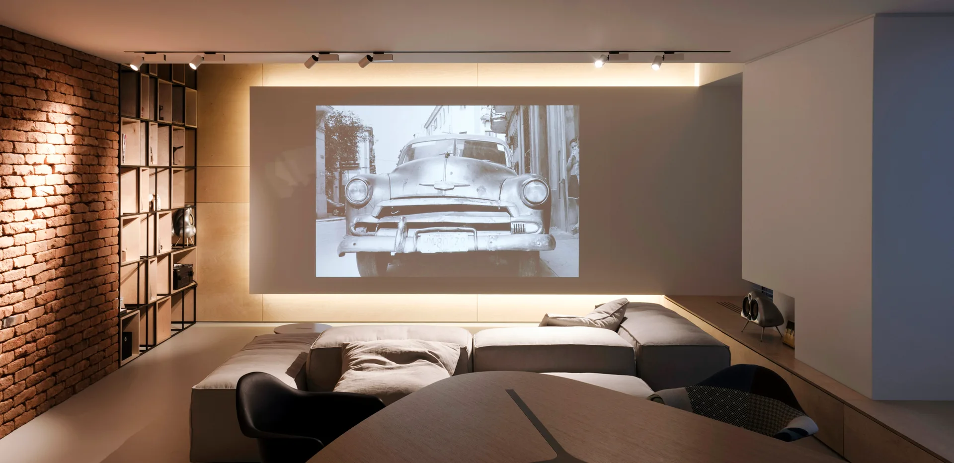 A modern home theater features a modular grey sofa facing a large wall-mounted projector screen, flanked by an exposed brick wall and grid shelving.