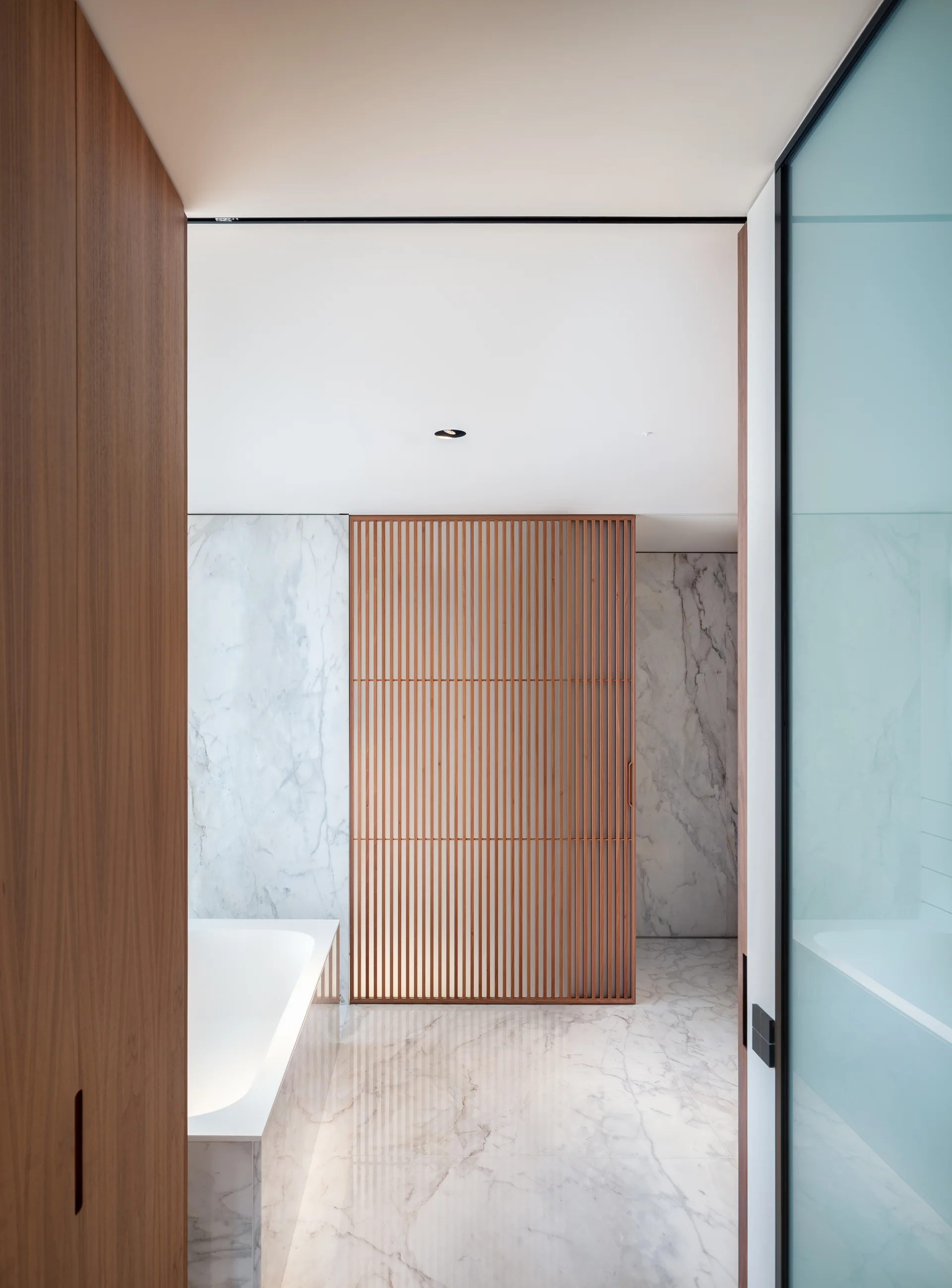 A modern bathroom interior showing a white marble bathtub, marble tiled floors and walls, and a tall vertical wood slat partition screen.