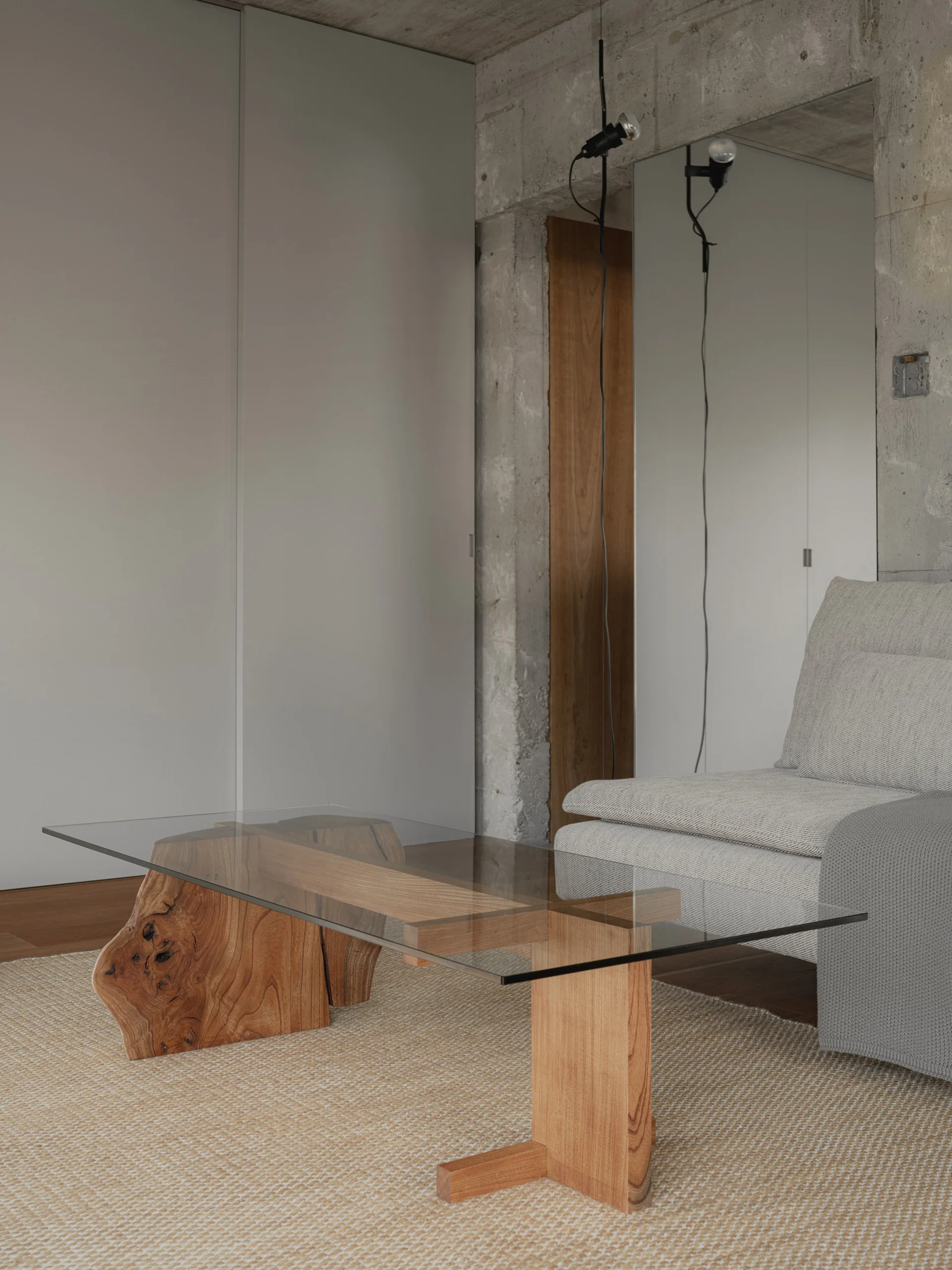 A minimalist living room features a glass-topped coffee table with a live-edge wood base on a woven rug near a concrete wall.