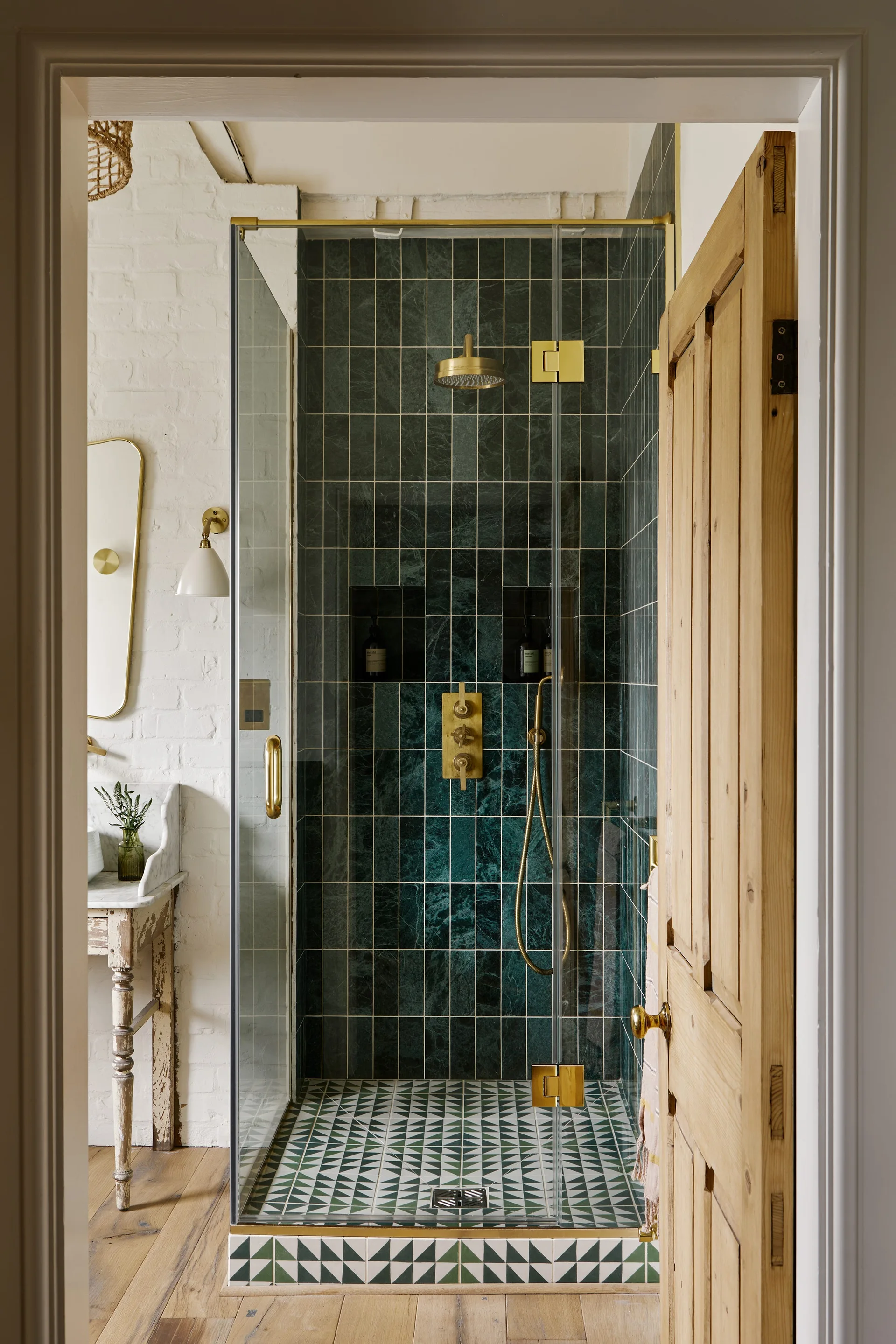 A bathroom features a glass shower with emerald green tiles, brass fixtures, and geometric floor tile next to a rustic wood vanity against painted brick.